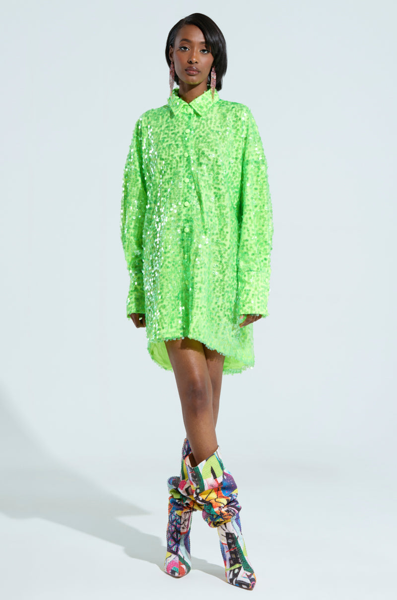 The individual poses in front of a simple backdrop, showcasing the vibrant VERONICA SEQUIN COLLARED SHIRT DRESS, complemented by eye-catching, patterned knee-high boots. Their shoulder-length dark hair enhances the look, while long, dangling earrings add a dynamic touch with each motion.