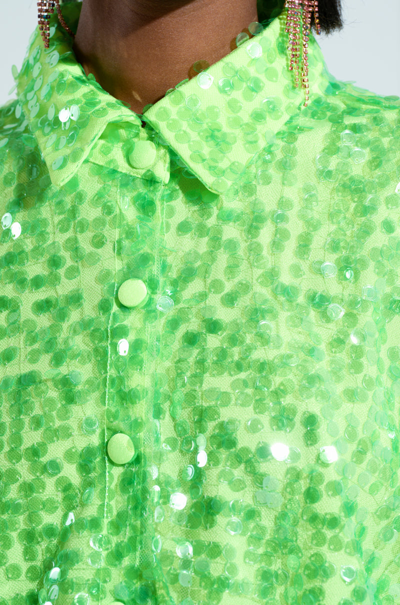A close-up of someone wearing a vibrant green oversized-fit VERONICA SEQUIN COLLARED SHIRT DRESS is shown, featuring large buttons and shimmering sequins. Partially visible are long, dangling earrings that gracefully complement the dress.