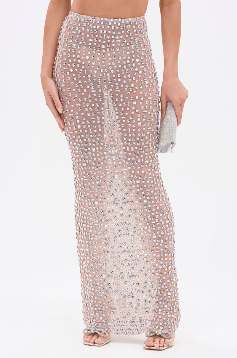 A person in the PRETTY LIKE ME RHINESTONE MESH MAXI SKIRT—a sheer, high-rise, floor-length style with sparkling rhinestones—stands barefoot and holding a silver clutch against a plain white background.