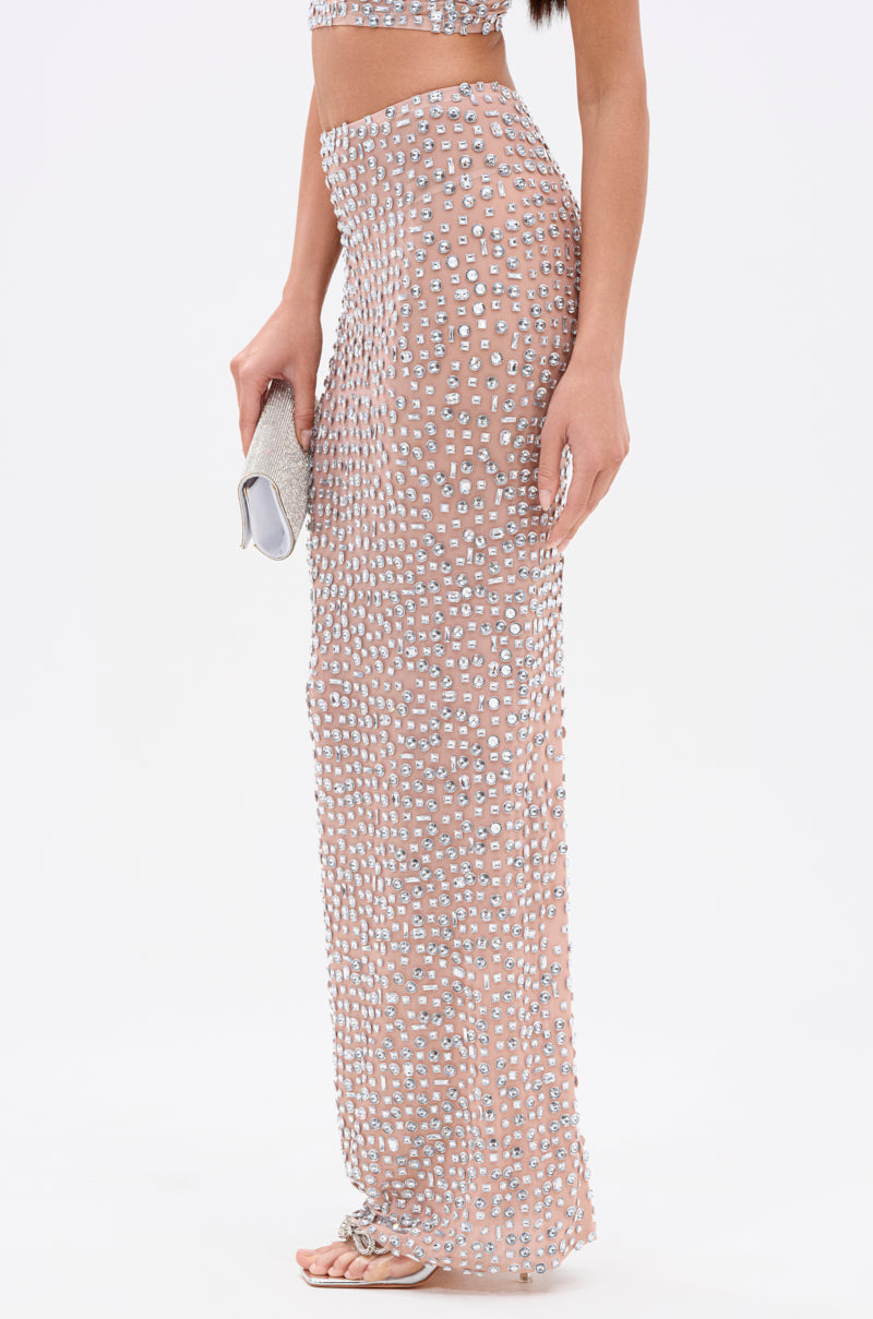 A woman models the PRETTY LIKE ME RHINESTONE MESH MAXI SKIRT, a sheer, high-rise maxi adorned with silver rhinestones. She carries a silver clutch and wears flat sandals. Only her torso and lower body appear against a white background.