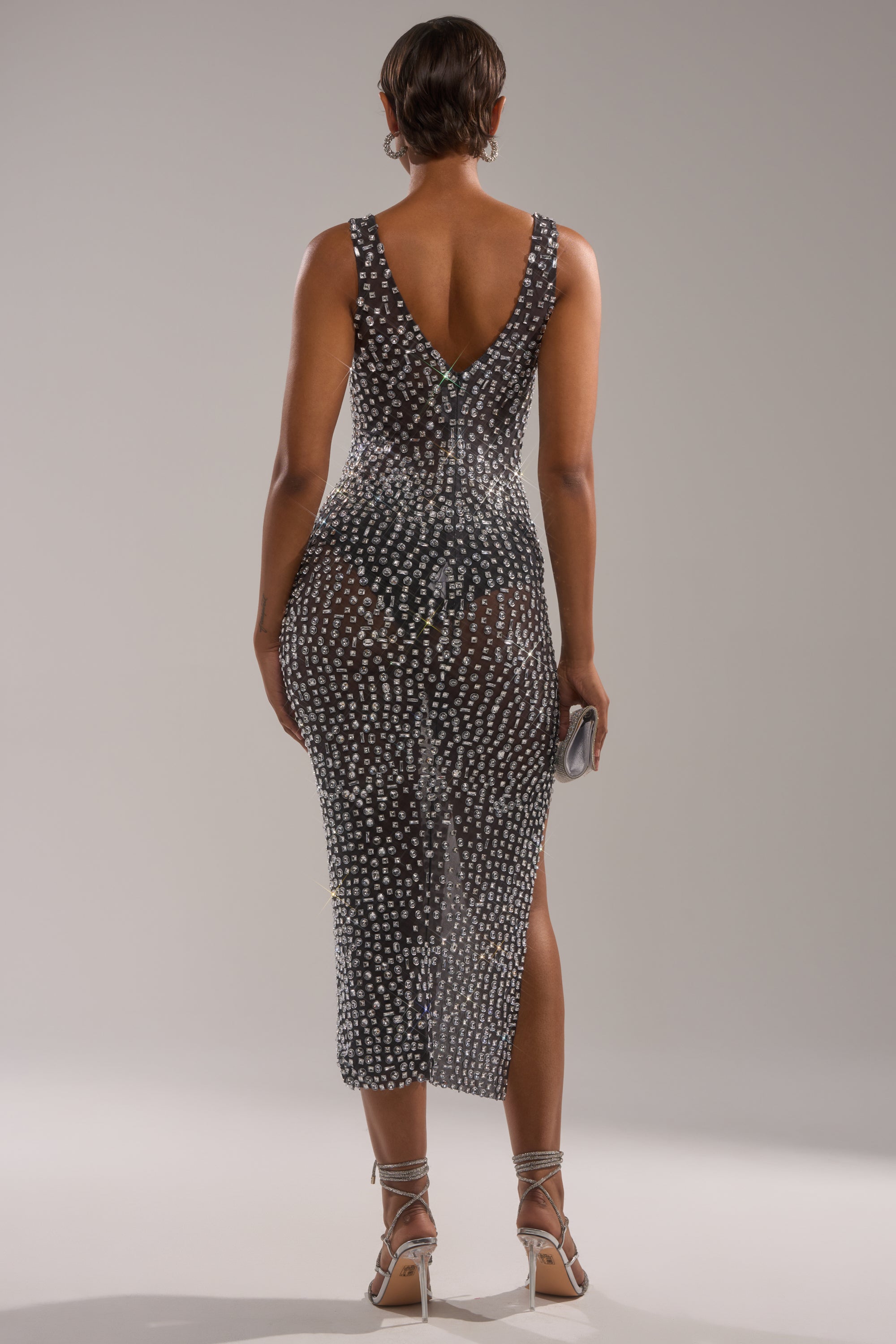A person wears the ROXANNE RHINESTONE STUDDED MAXI DRESS IN BLACK, a curve-hugging sleeveless mesh dress with white dots, a deep V back, and below-the-knee hem. Styled with silver high heels and a small clutch.