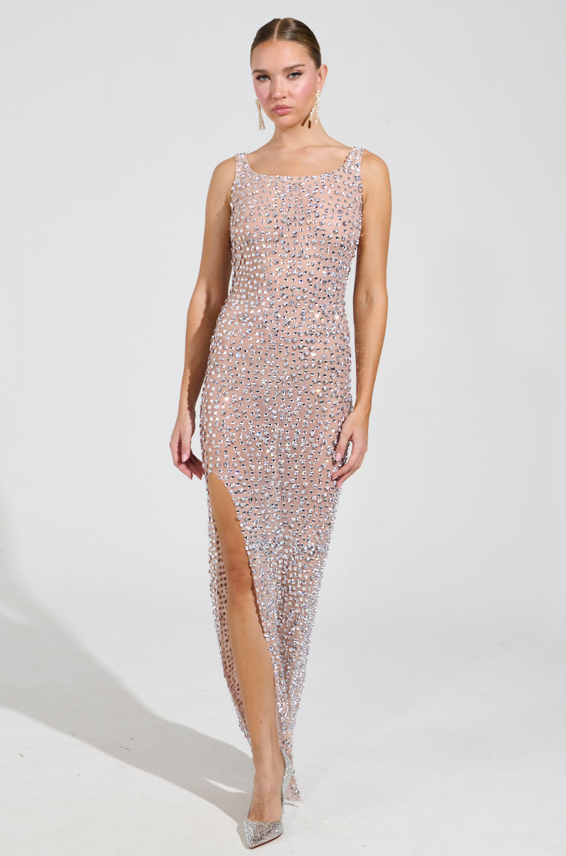 In a dazzling ROXANNE RHINESTONE STUDDED MAXI DRESS, a woman stands confidently. Her sleeveless gown, adorned with sequins and featuring a high slit, shimmers under the light. Her hair is styled back and paired with pointed heels against a plain, light-colored background.