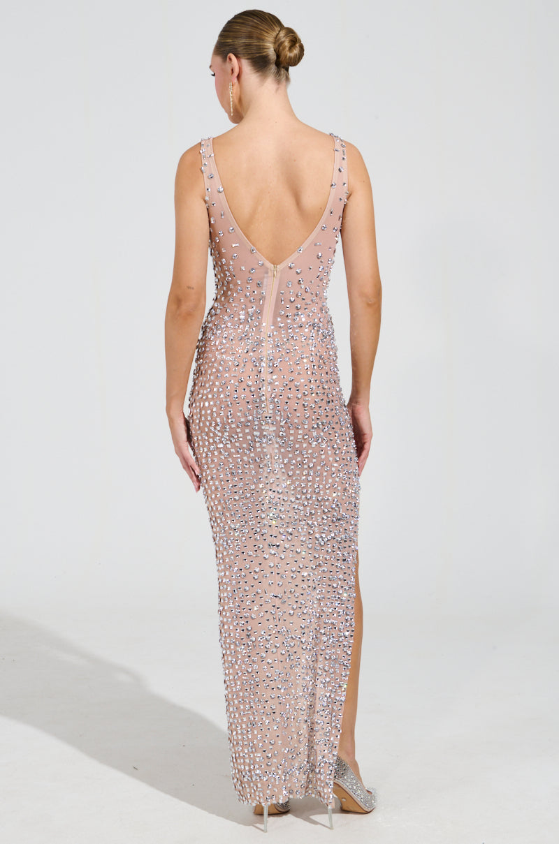 The individual is wearing the dazzling ROXANNE RHINESTONE STUDDED MAXI DRESS, characterized by its sleeveless design, low back, and intricate beadwork. They are facing away with their hair neatly styled in a bun. This semi-transparent gown is adorned with silver rhinestones and includes a back slit for an extra touch of elegance.