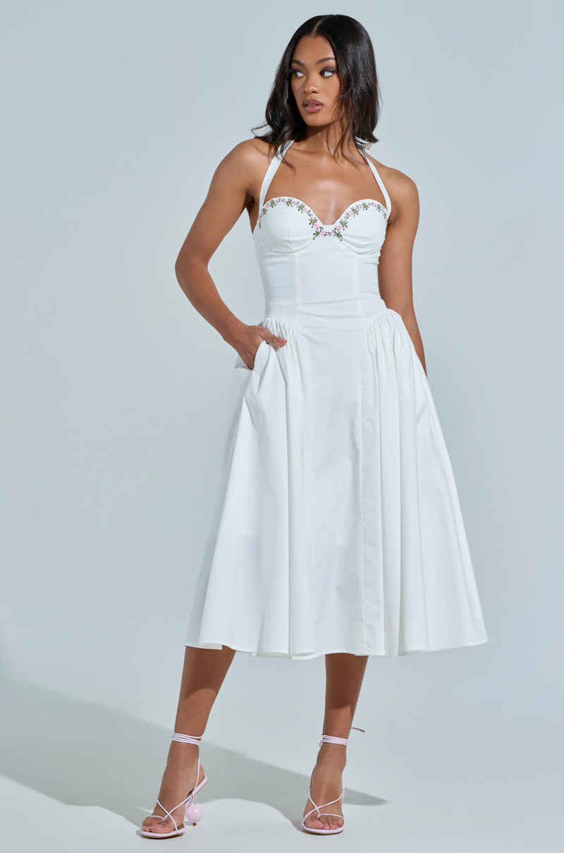 A model wears the "Off to Brunch" poplin midi dress, featuring a white halter-neck and corset-like bodice with floral embroidery, posing with hands in pockets against a light gray background. She completes the look with strappy high heels.