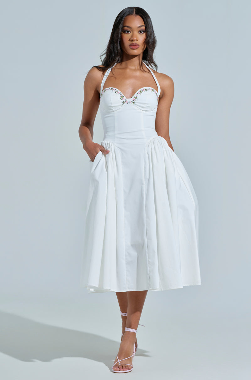 A woman poses in the OFF TO BRUNCH POPLIN MIDI DRESS, featuring a white halter-neck, floral embroidery, corset-like bodice, full skirt with pockets. She completes the look with strappy high-heeled sandals against a plain background.