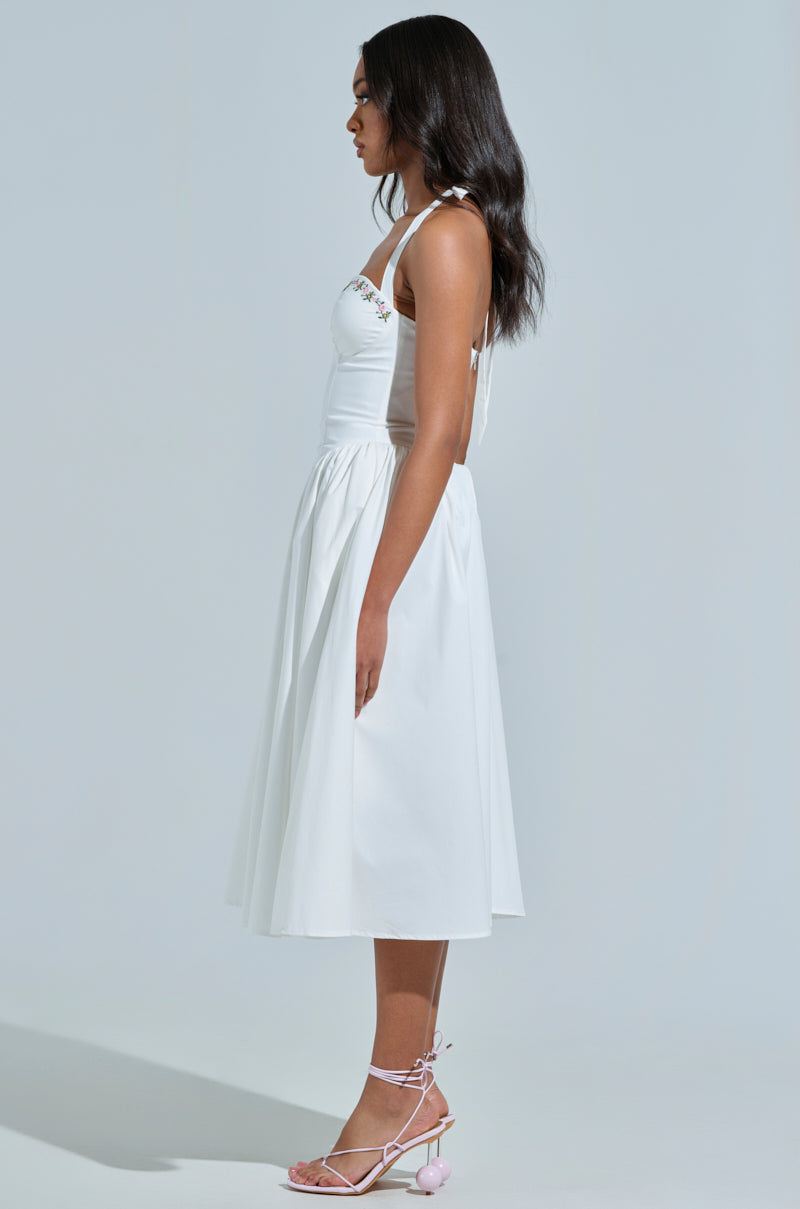 A person with long hair stands in profile wearing the OFF TO BRUNCH poplin midi dress featuring thin straps and a corset-like bodice. Their sandals have decorative pom-poms. The background is a plain, neutral color.