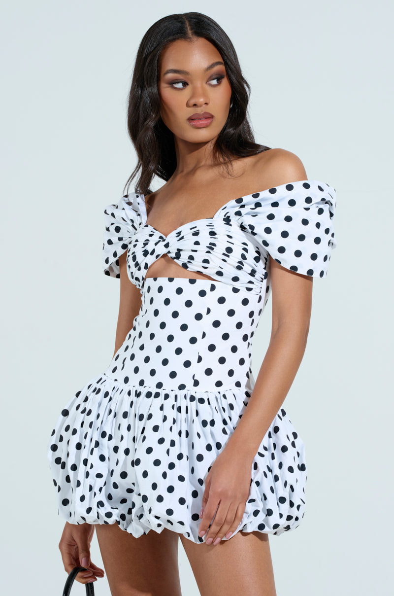 A woman, wearing the 'Cutest One at the Function' Polka Dot Mini Dress with a twisted front, puffed short sleeves, voluminous skirt, and sweetheart neckline, poses against a plain light background. Her long wavy hair adds to the elegant look.