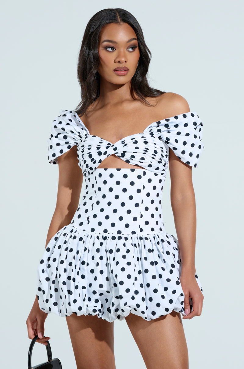 A woman wears the CUTEST ONE AT THE FUNCTION POLKA DOT MINI DRESS, showcasing its off-the-shoulder puff-sleeve and front cutout. She holds a small black handbag, her dark hair in loose waves against a plain light blue background.