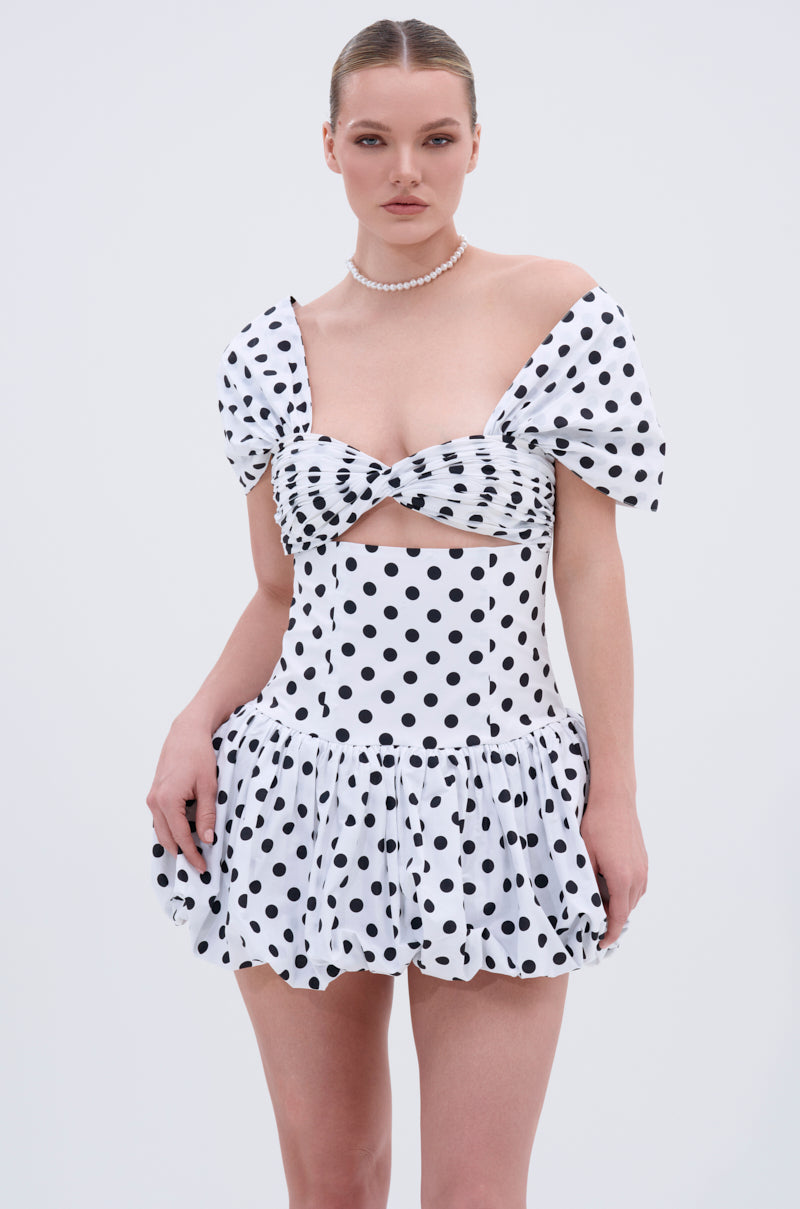 A woman wears the CUTEST ONE AT THE FUNCTION POLKA DOT MINI DRESS featuring off-the-shoulder puffy sleeves, a sweetheart neckline, and a bubble skirt. She accessorizes with a pearl necklace and has slicked-back hair against a plain light background.