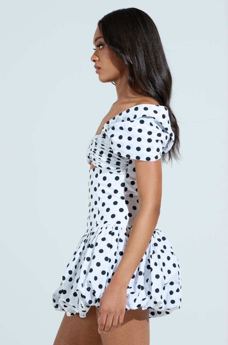 A woman stands in profile wearing the CUTEST ONE AT THE FUNCTION POLKA DOT MINI DRESS, enhancing her graceful silhouette with its off-the-shoulder puff sleeves and fitted bodice. Her long dark hair cascades down against a plain background, perfectly complementing the flared skirt of the dress.