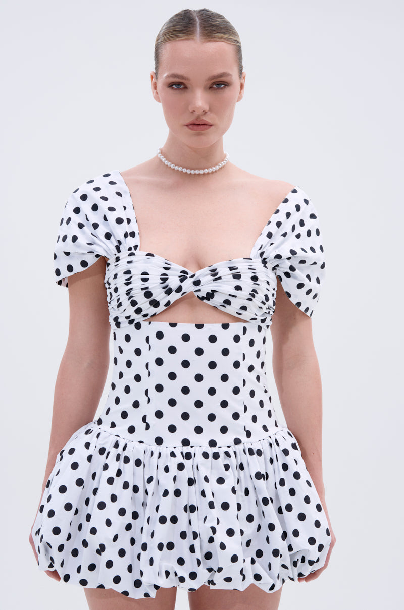 A woman wears the CUTEST ONE AT THE FUNCTION Polka Dot Mini Dress, featuring puffed short sleeves, a front cutout, and a ruched skirt. Her hair is slicked back and she accessorizes with a pearl necklace. The background is light gray.