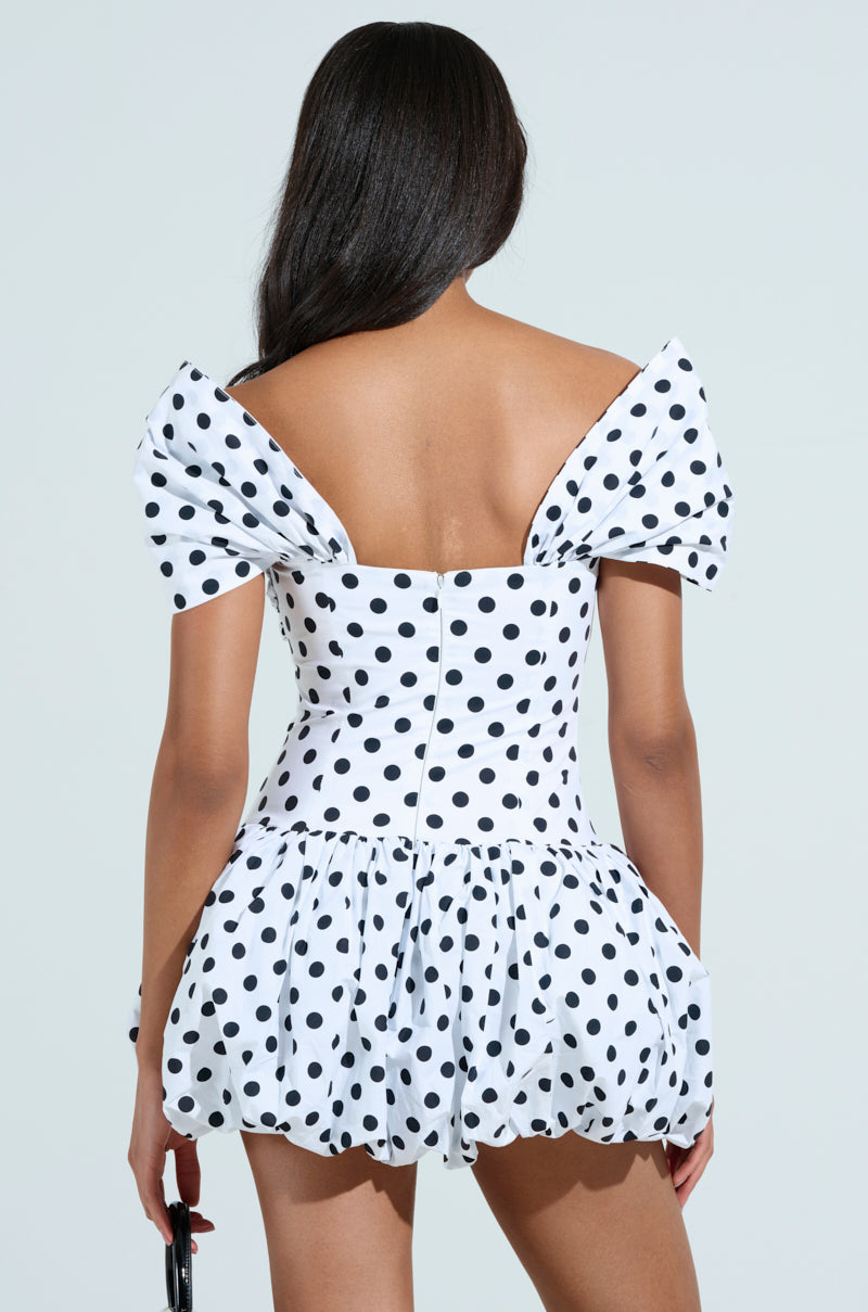 Rear view of a person with long dark hair wearing the CUTEST ONE AT THE FUNCTION POLKA DOT MINI DRESS, an off-the-shoulder mini dress in white with black dots, fitted bodice, flared skirt, and puffed sleeves set against a plain light blue background.