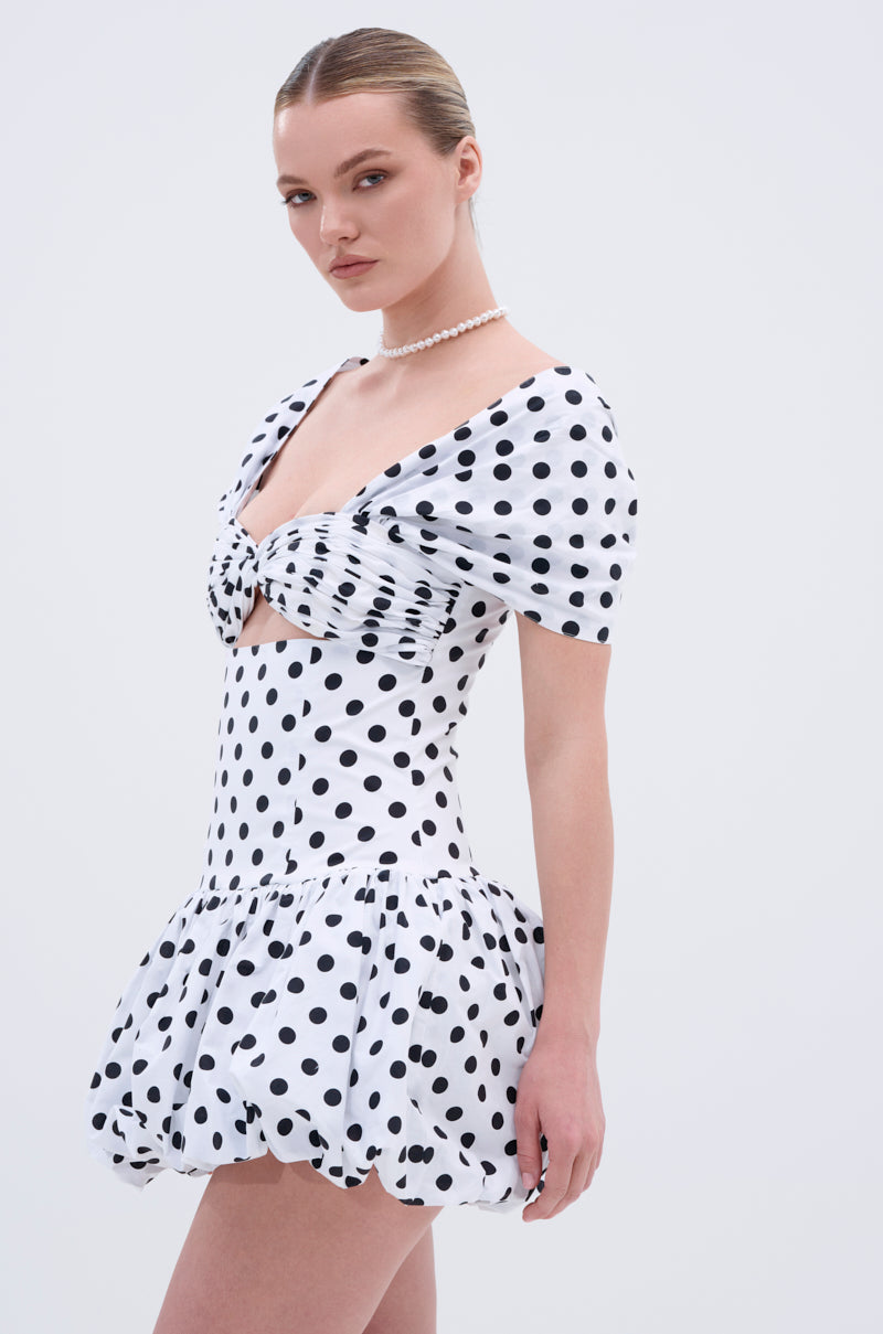 A woman wears the CUTEST ONE AT THE FUNCTION POLKA DOT MINI DRESS, featuring puff sleeves, a ruched front cut-out, bubble hem, and sweetheart neckline. Her hair is tied back and she accessorizes with a pearl necklace against a plain background.