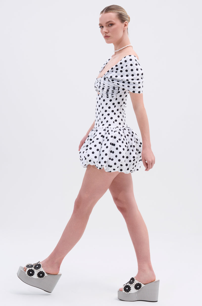 A person in the CUTEST ONE AT THE FUNCTION POLKA DOT MINI DRESS, a white off-the-shoulder mini with puffed sleeves, walks sideways. They have blonde hair in a low ponytail and wear daisy platform sandals against a plain white background.