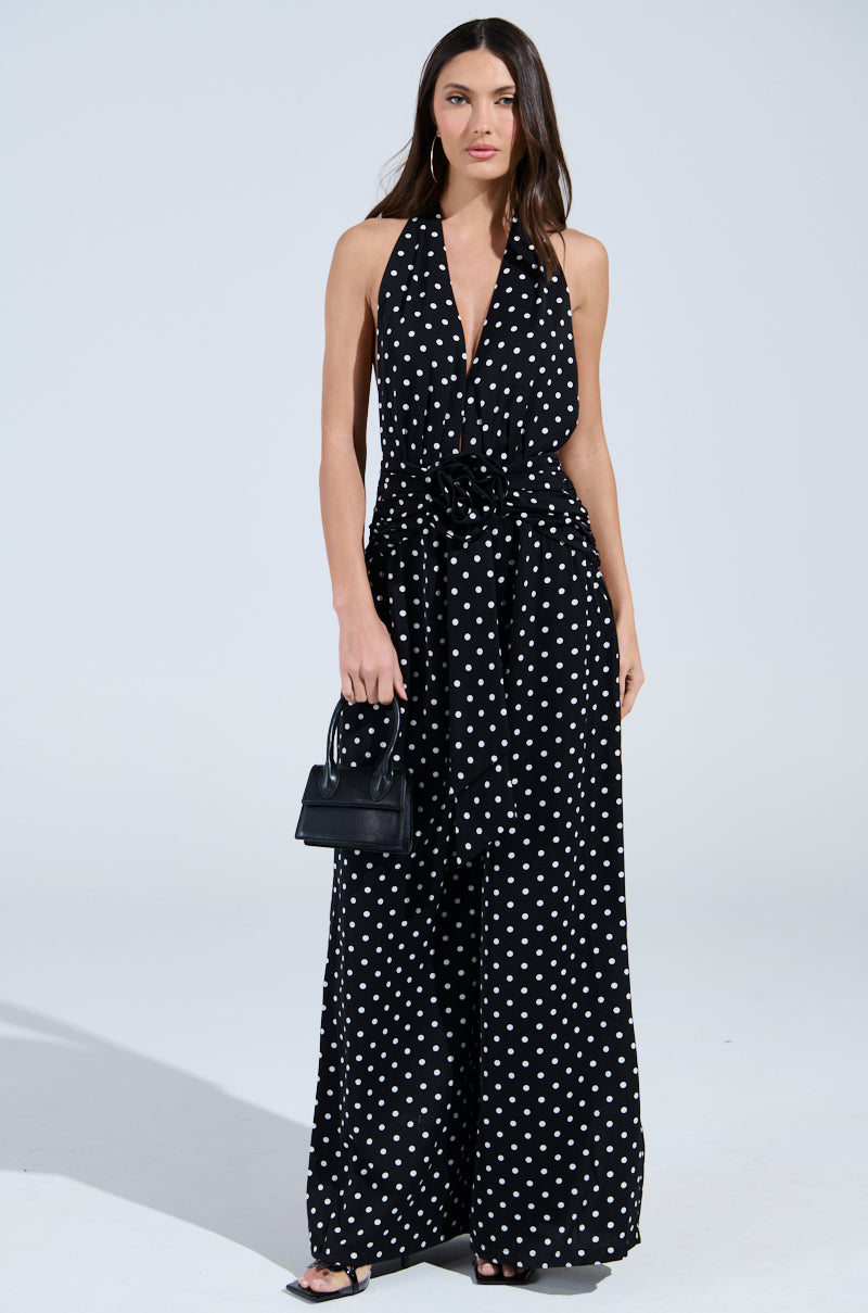 A woman models the BEST DRESSED POLKA DOT JUMPSUIT with a deep V-neckline and wide-leg silhouette. She completes her chic look with a black handbag and heels, standing against a simple light backdrop.