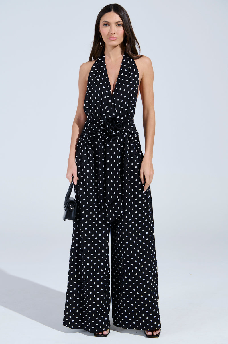 A woman wears the BEST DRESSED POLKA DOT JUMPSUIT—a retro-chic, sleeveless black jumpsuit with wide-leg pants and a tied waist belt—paired with a small black handbag against a plain light background.