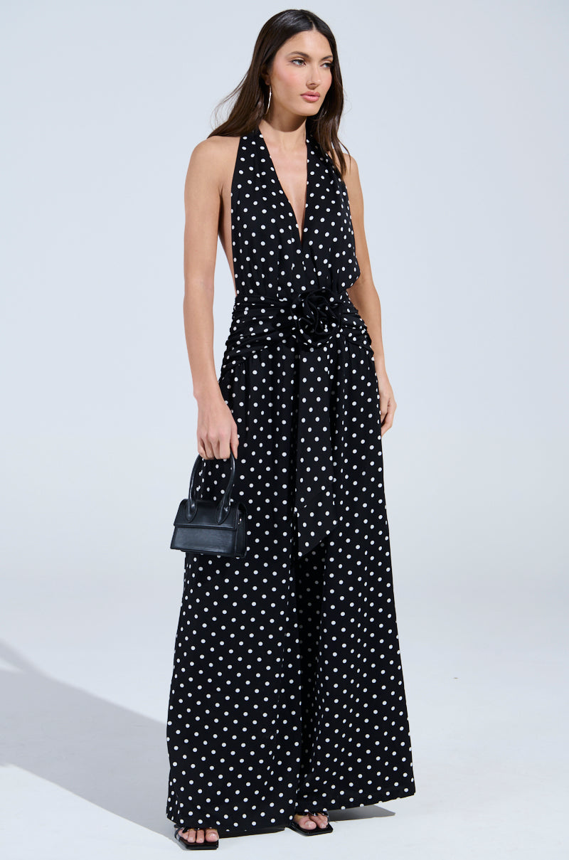 A woman models the BEST DRESSED POLKA DOT JUMPSUIT, featuring a deep V-neckline, wide tie belt, and wide-leg silhouette. She pairs it with a black handbag and open-toe heels against a simple light backdrop.