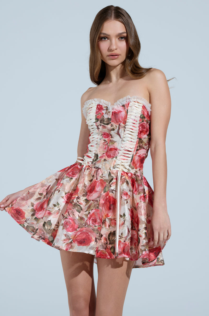 Against a light blue backdrop, a woman dons the KEEP DREAMIN FLORAL MINI DRESS. This strapless design features a ruffled bust and flared skirt, decorated with red and pink roses on a light background, creating an elegant silhouette.