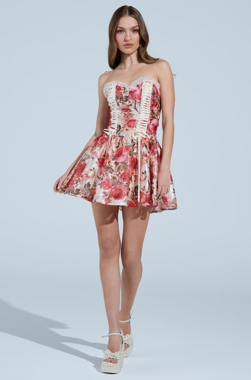 Wearing the KEEP DREAMIN FLORAL MINI DRESS, showcasing pink and red roses with a lace-up front, a woman stands against a light blue backdrop. Her strapless silhouette highlights her long, loose hair, complemented by white heeled sandals.