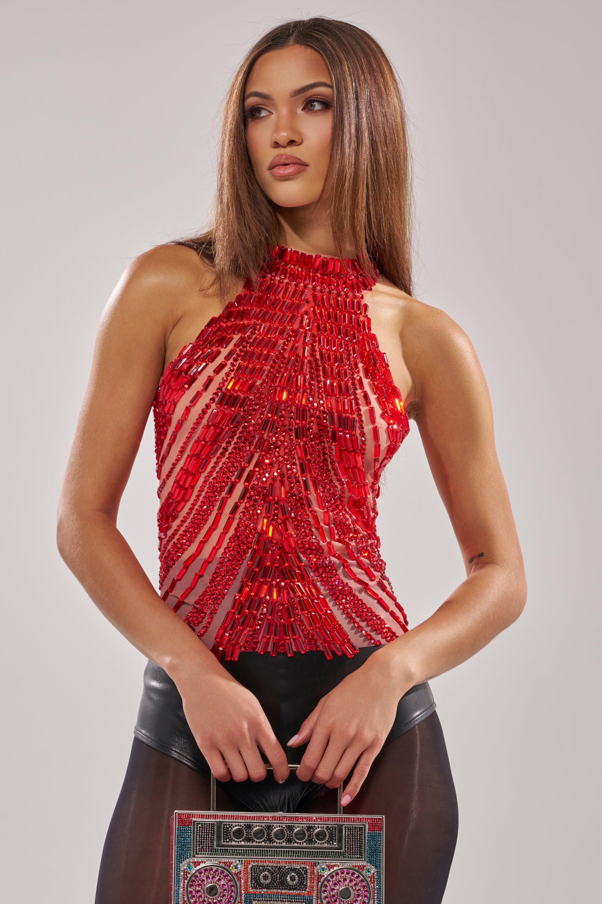 A woman with straight brown hair wears the FIRECRACKER RHINESTONE BLOUSE, a red sleeveless top with beads and sheer fabric, paired with high-waisted black pants. She holds a colorful clutch bag and gazes to the side.