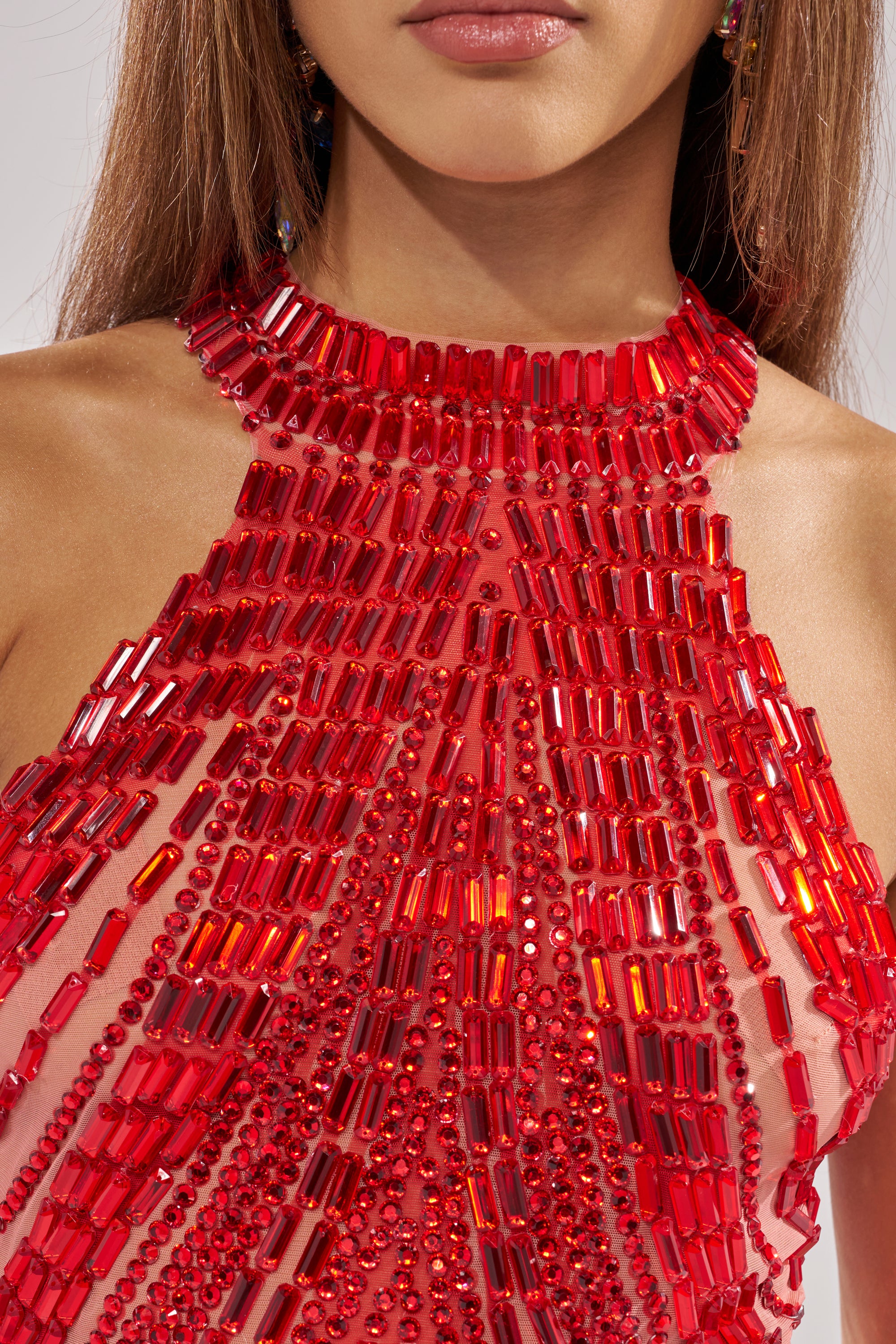 A close-up of a woman wearing the FIRECRACKER RHINESTONE BLOUSE, a cropped sleeveless top featuring intricate red rhinestones and geometric rectangular details on sheer fabric. Her straight brown hair partly covers her face, showing only the lower part.