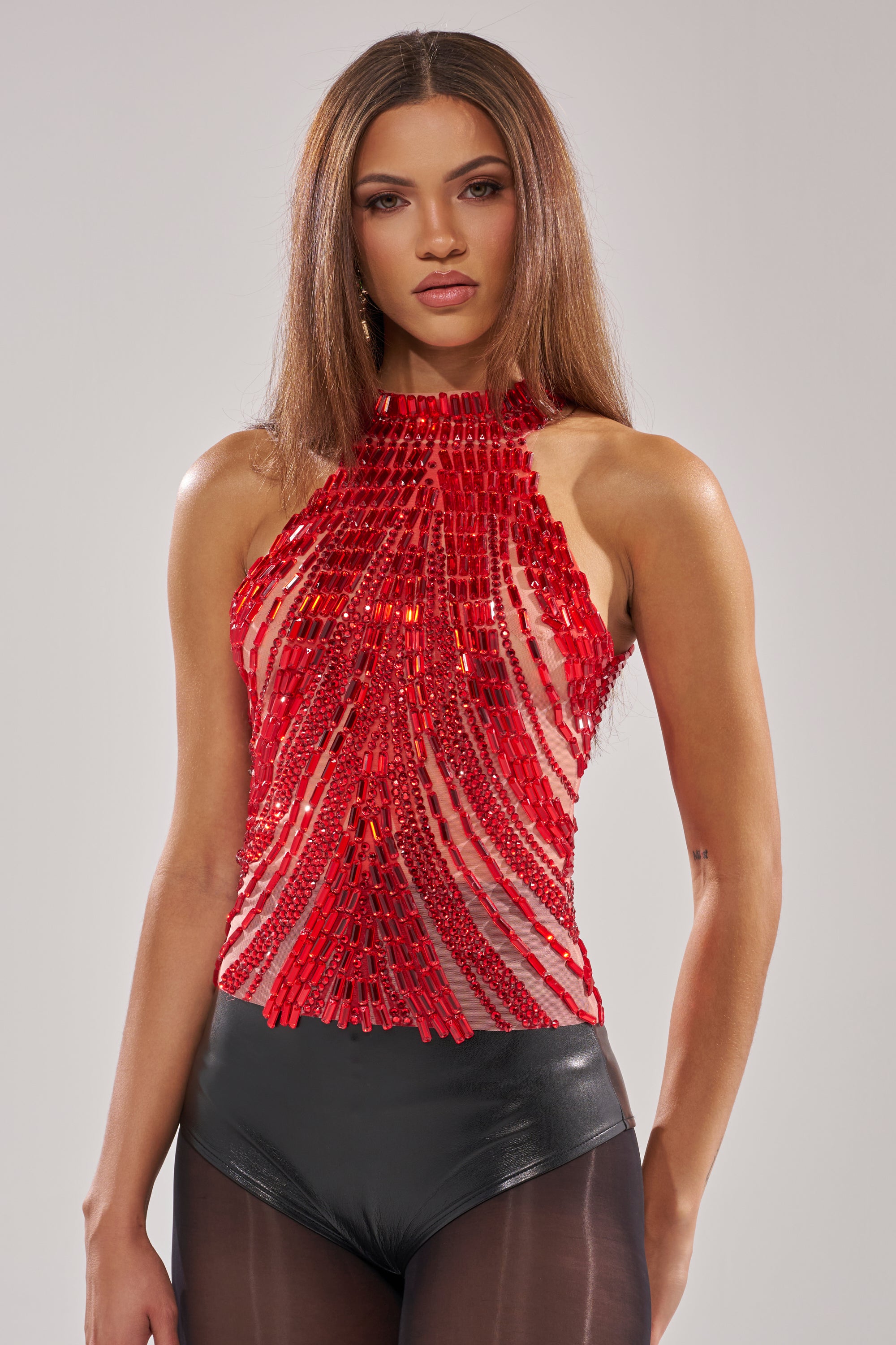 A woman with long straight hair wears the FIRECRACKER RHINESTONE BLOUSE—cropped, sleeveless, and adorned with geometric rhinestone patterns—paired with black high-waisted bottoms and sheer tights, posing against a plain light background.