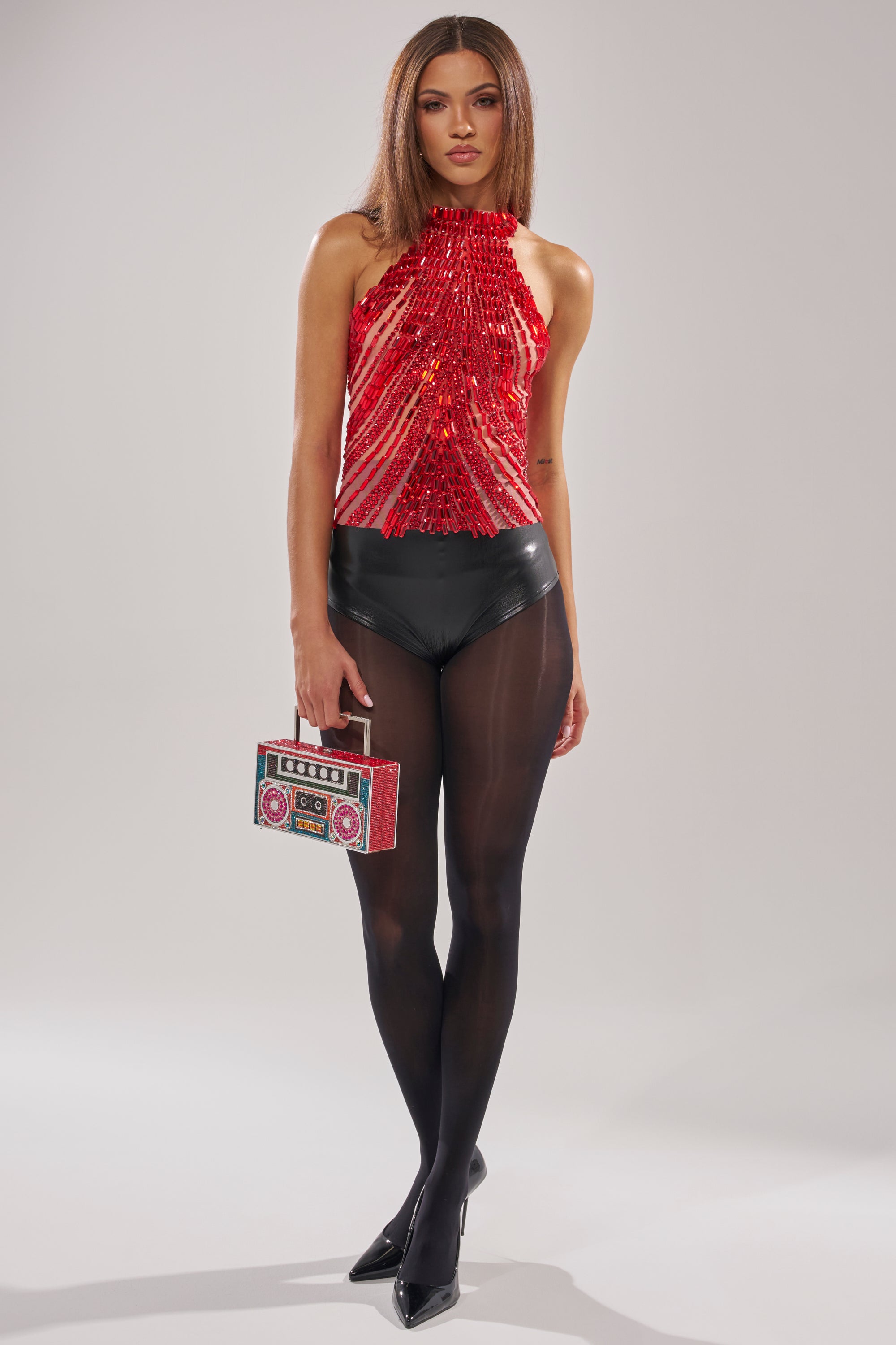 A woman with straight brown hair and a neutral expression wears the FIRECRACKER RHINESTONE BLOUSE, black high-waisted shorts, black sheer tights, and heels, carrying a retro boombox-shaped clutch.