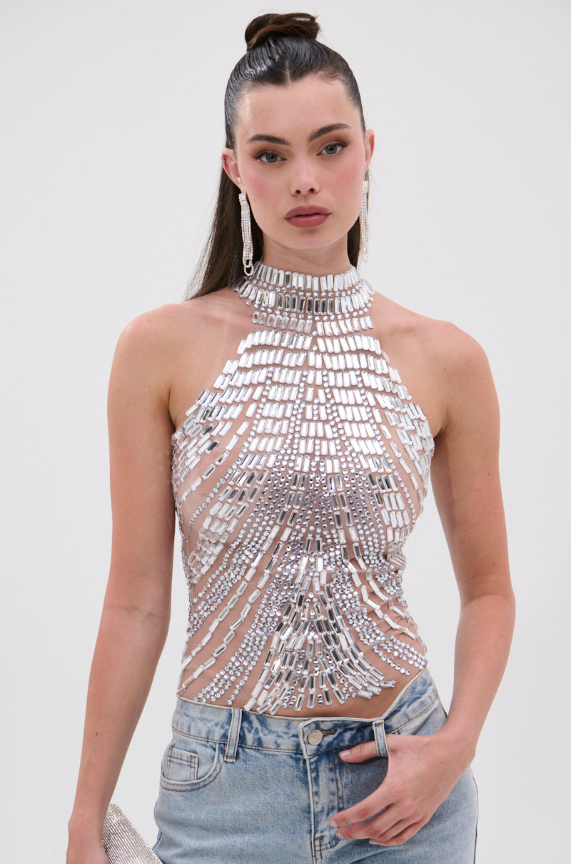 A woman wears the YOU KNOW THE DRILL RHINESTONE BLOUSE, a sleeveless, high-neck silver mirrored rhinestone top, with light blue jeans. Her long dark hair is styled in a high ponytail and she holds a small silver clutch.