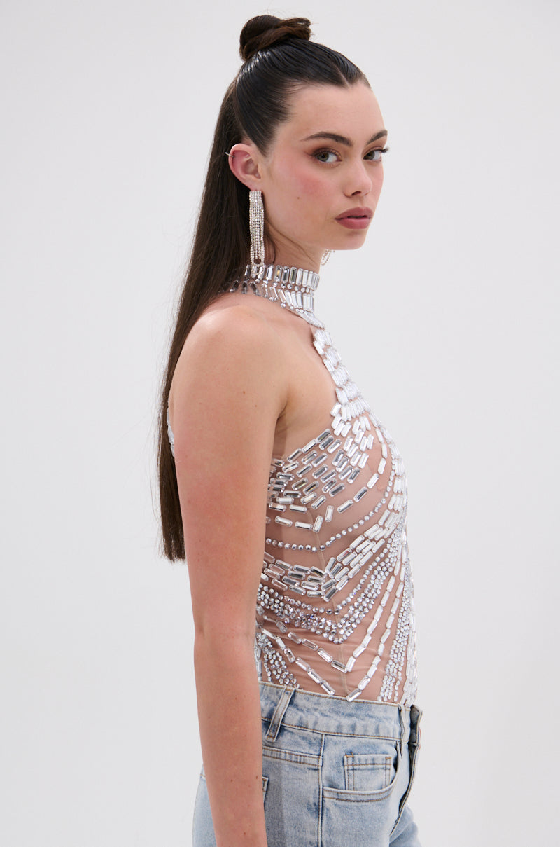 A woman with long brown hair in a high ponytail wears the YOU KNOW THE DRILL RHINESTONE BLOUSE—a sheer, sleeveless, high-neck top with silver sequin details—paired with light blue jeans and dramatic earrings, highlighting the chic cropped fit.