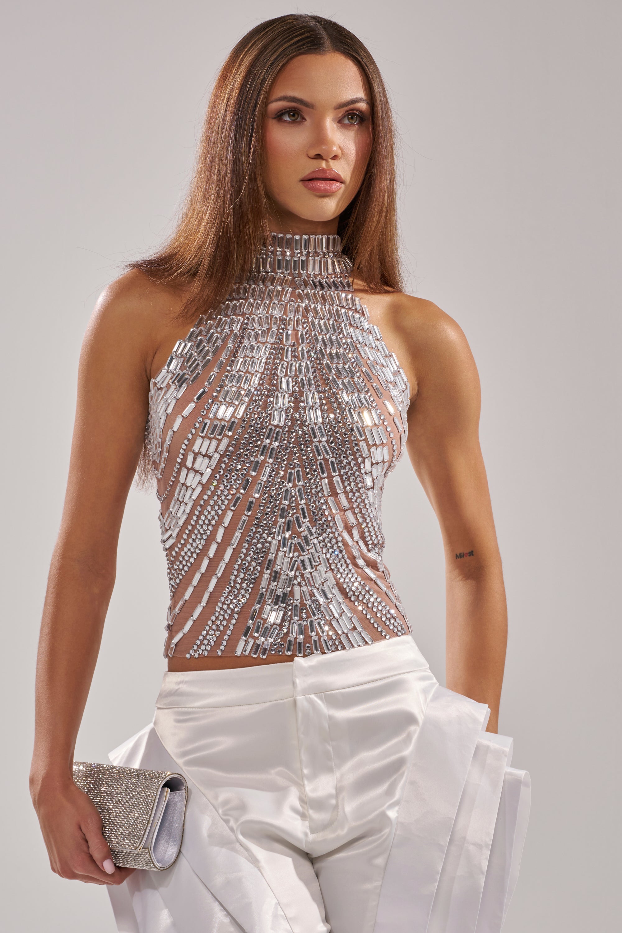 A woman stands against a plain background wearing the YOU KNOW THE DRILL RHINESTONE BLOUSE, a cropped sleeveless top adorned with reflective silver rhinestones, paired with white satin pants and holding a glittery silver clutch.