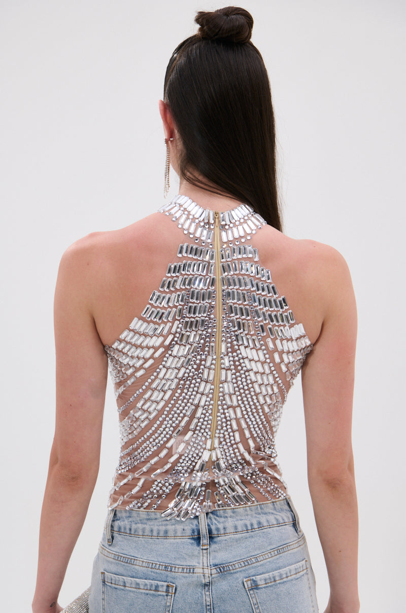 A person with long, straight brown hair in a topknot wears the YOU KNOW THE DRILL RHINESTONE BLOUSE—sleeveless, sheer mesh with silver and white beadwork and a visible back zipper—paired with light blue jeans.