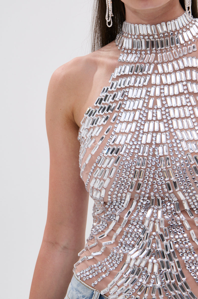 A woman wears the YOU KNOW THE DRILL RHINESTONE BLOUSE—sleeveless, high-neck, adorned with silver and mirrored beads—paired with light blue denim jeans. Only her torso and lower face are shown.