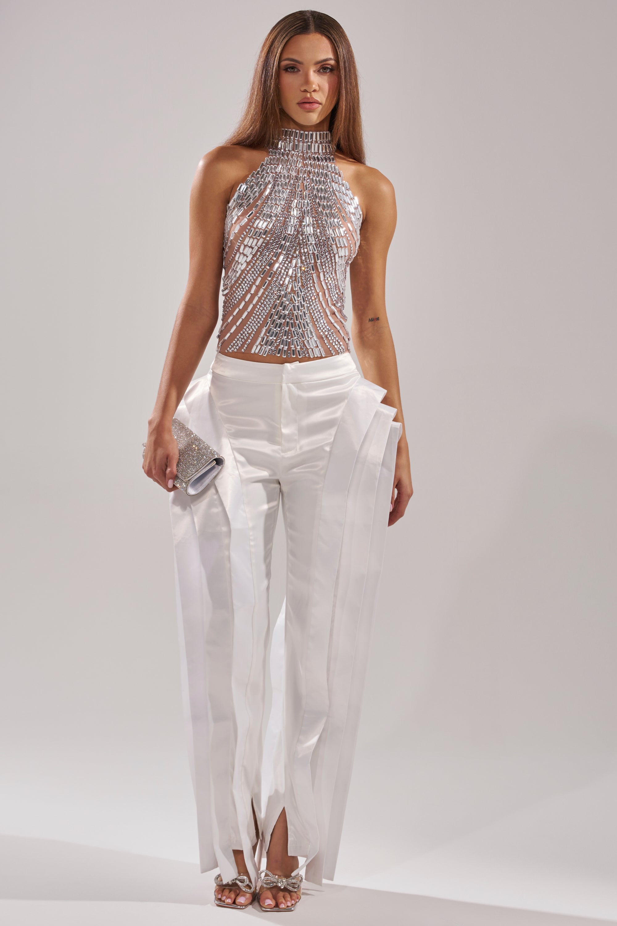 A woman stands against a plain background wearing the YOU KNOW THE DRILL RHINESTONE BLOUSE with high-waisted white satin pants featuring long vertical strips. She accessorizes with a silver clutch and strappy heels.