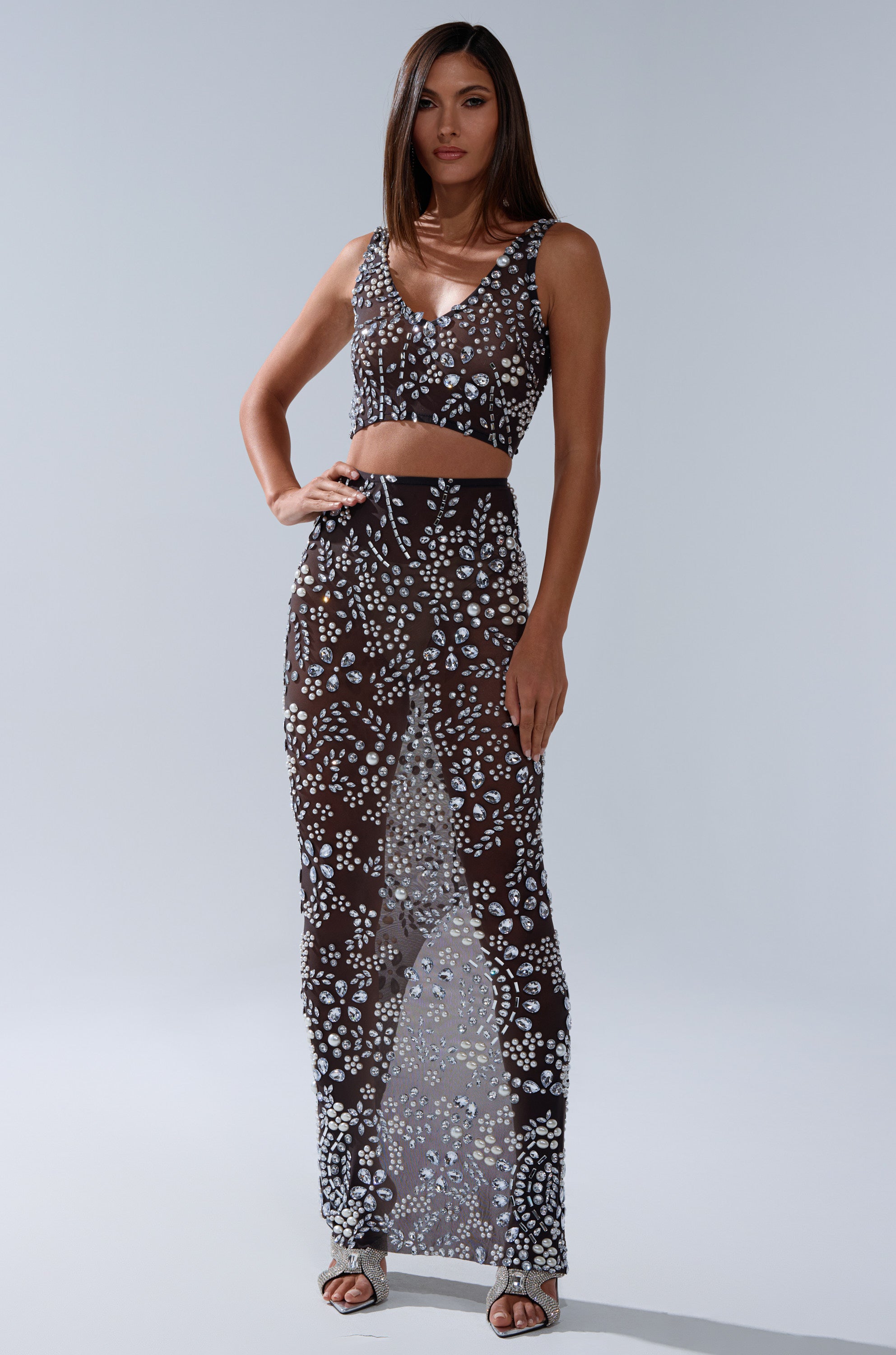 A woman poses in the BLING BLING MESH RHINESTONE MAXI SKIRT, a sheer black high-rise mesh skirt with white rhinestone detailing. She pairs it with a matching crop top, strappy sandals, and her straight hair loose.