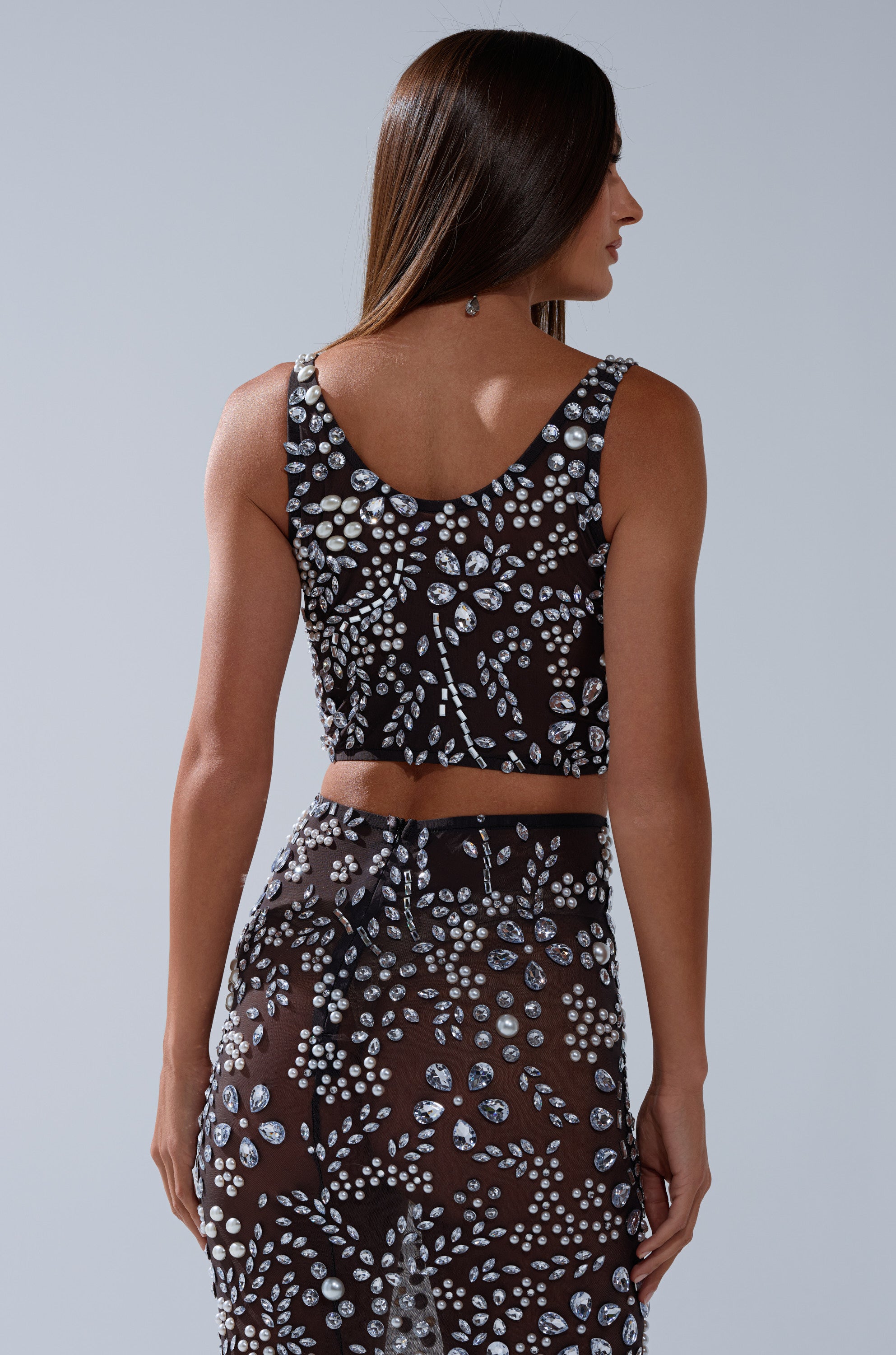 A woman with long brown hair faces away, wearing the BLING BLING MESH RHINESTONE BRALETTE in black with matching sheer, silver-sequined skirt, against a plain light gray background.