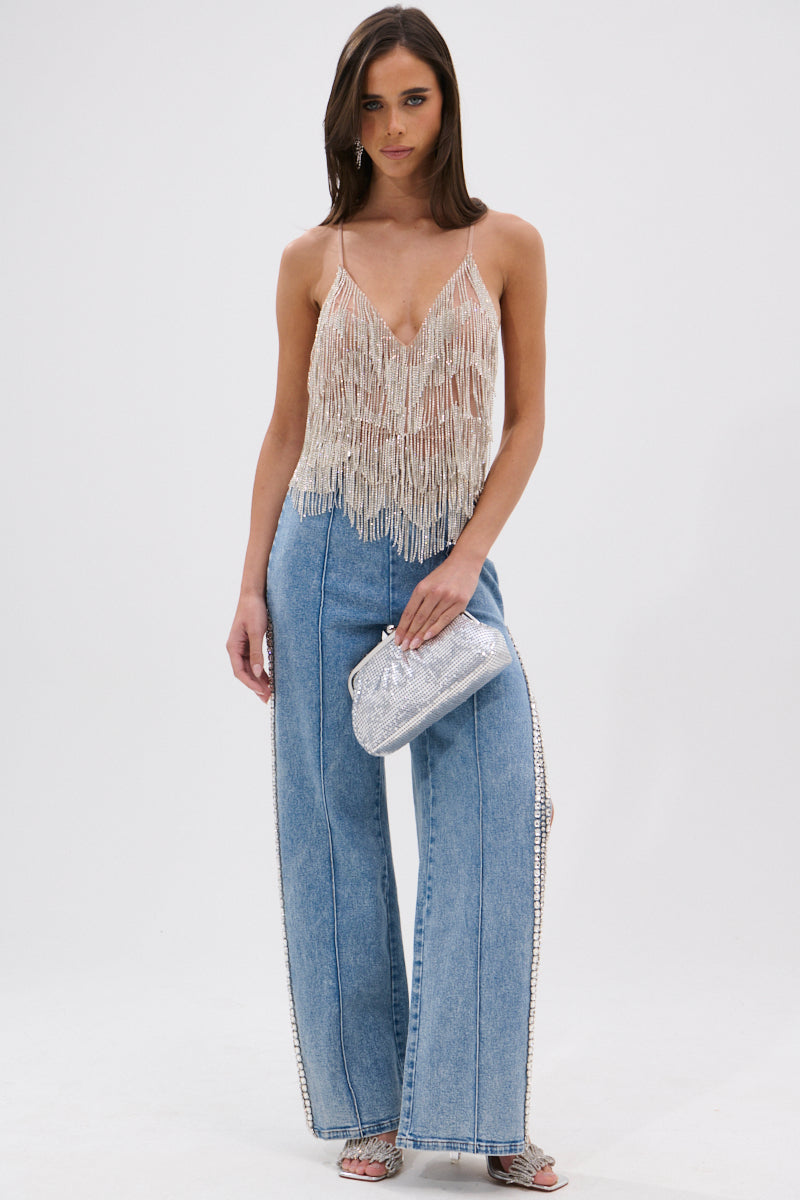 A woman stands against a plain background wearing the SHINING STAR RHINESTONE FRINGE TANK TOP with a plunging V-neckline and high-waisted wide-leg blue jeans, accessorized with a silver clutch and silver heels.