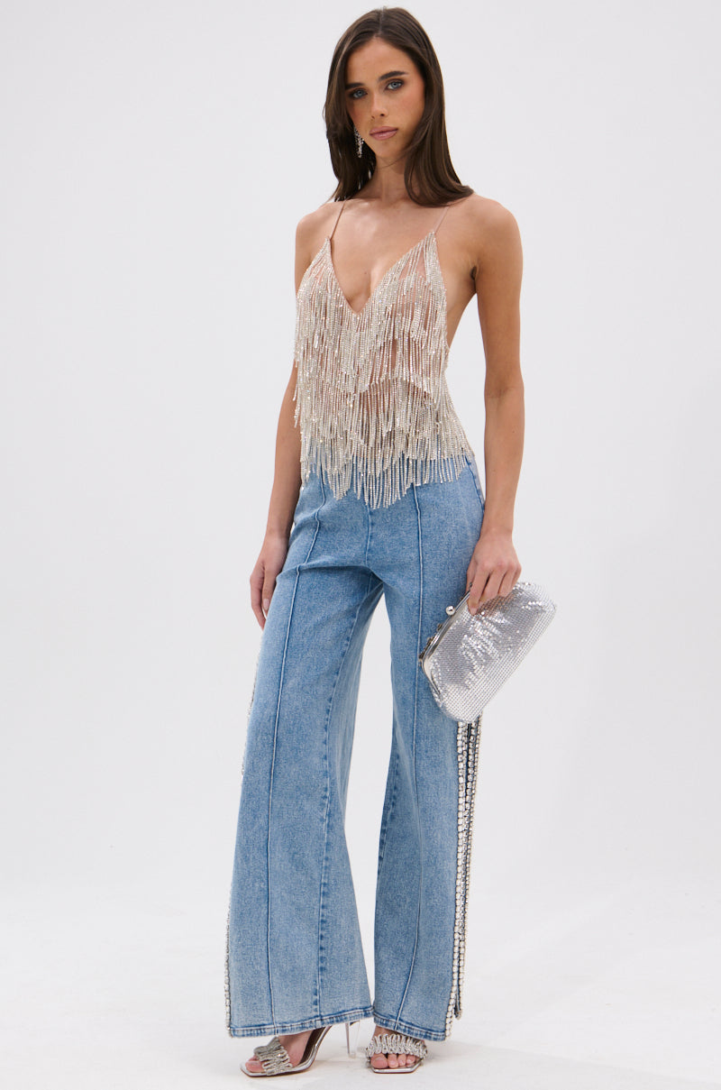 A woman poses against a white background wearing the SHINING STAR RHINESTONE FRINGE TANK TOP, paired with high-waisted wide-leg jeans, silver heeled sandals, and holding a silver sequin clutch.
