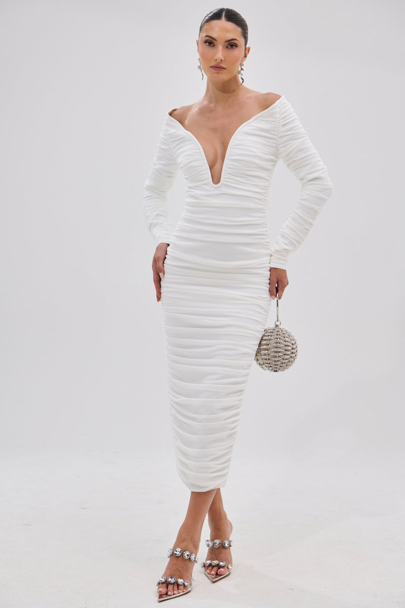 A woman wears the TARAJI Bandage Midi Dress with Mesh Overlay—fitted, long-sleeve, white with ruching and a deep V neckline—styled with a silver round handbag and embellished heeled sandals against a plain white background.