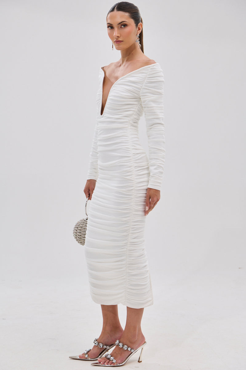 A woman poses against a plain white background in the TARAJI BANDAGE MIDI DRESS WITH MESH OVERLAY, featuring an off-the-shoulder design and deep V-neckline. She accessorizes with a round beaded purse and embellished pointed-toe heels.