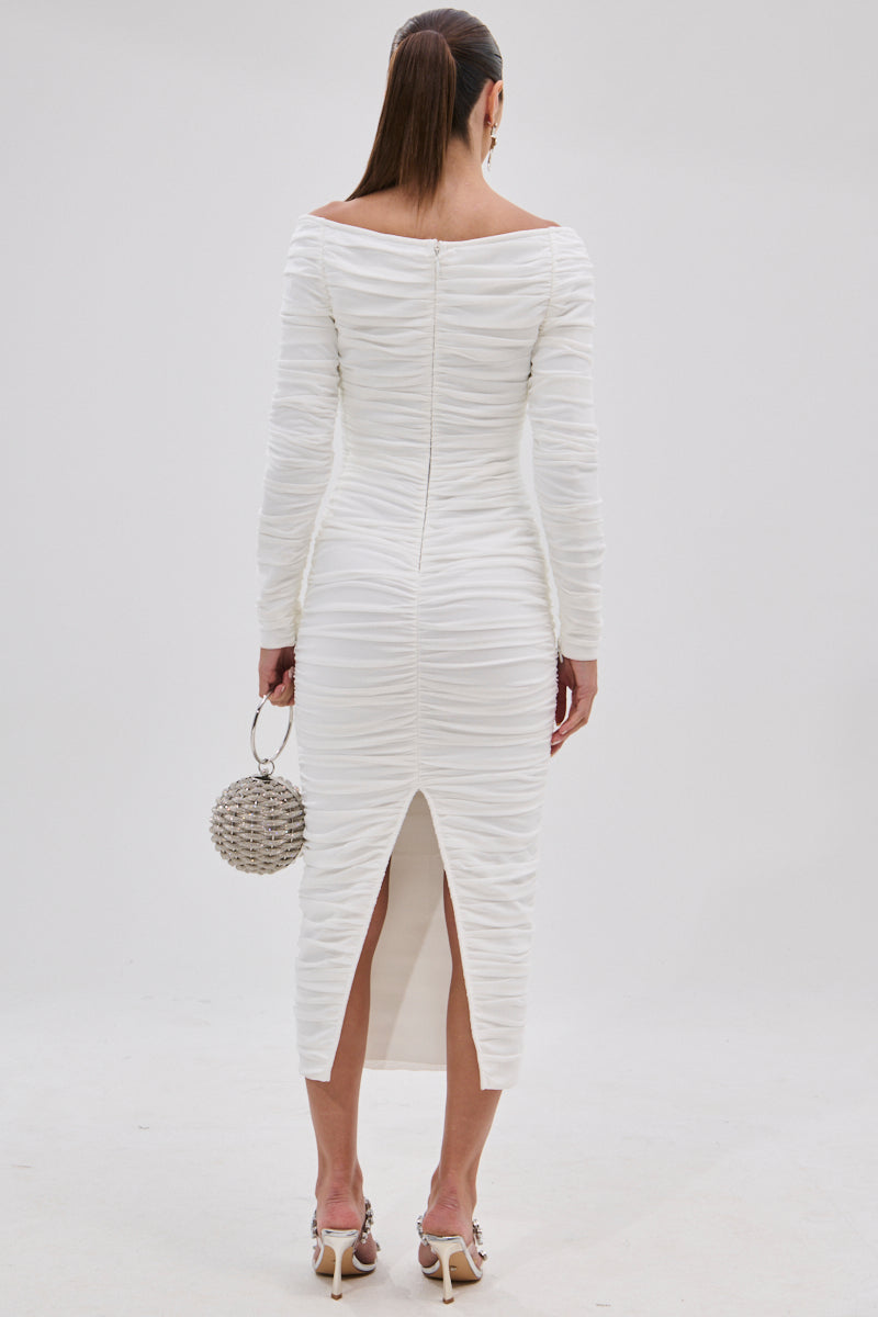A woman in the TARAJI Bandage Midi Dress with Mesh Overlay stands facing away. She holds a round beaded purse, wears heeled sandals, and her hair is styled in a sleek ponytail. The dress includes a back slit for added allure.