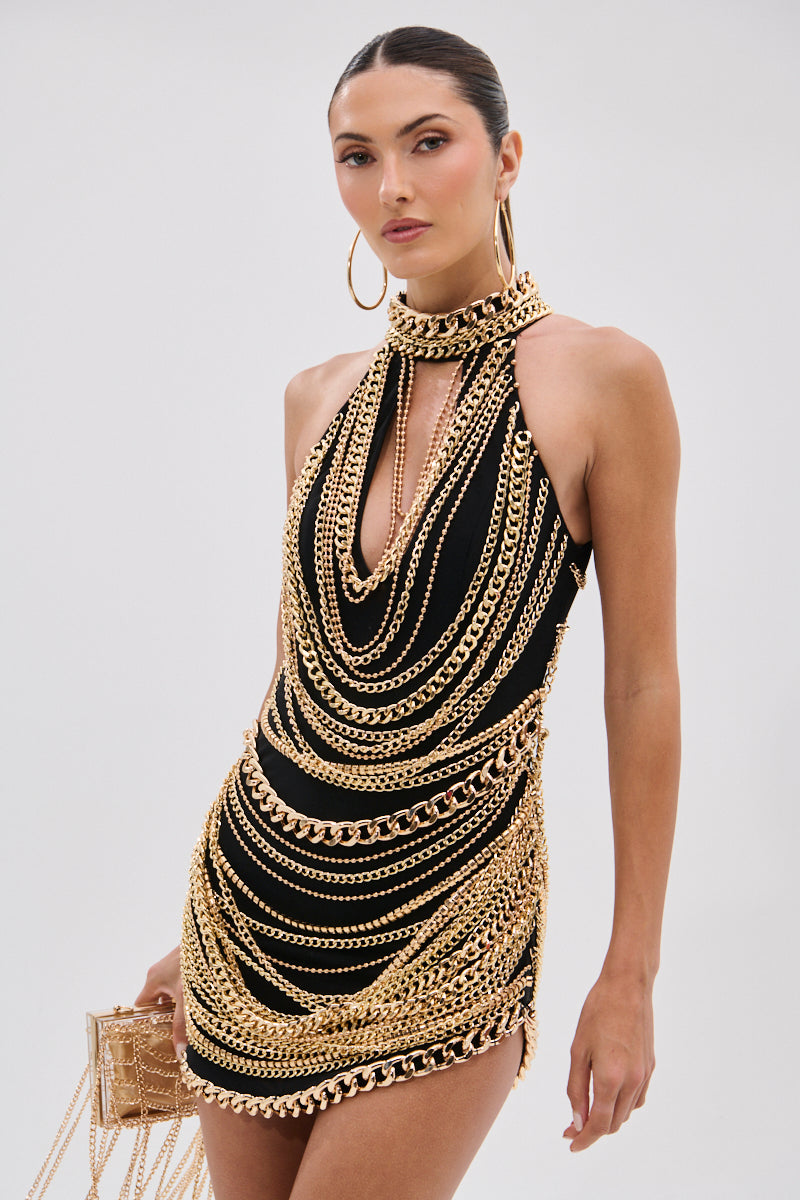 A woman wears the STAND OUT CHAIN MINI DRESS—sleeveless and mesh—with layered gold-tone chains, large hoop earrings, and a gold chain handbag, standing against a plain light background.