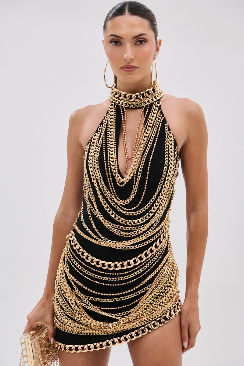 A woman models the STAND OUT CHAIN MINI DRESS, a sleeveless black mesh mini dress with bold gold-tone chains. She pairs it with a gold clutch and large gold hoop earrings, her hair slicked back, against a plain background.