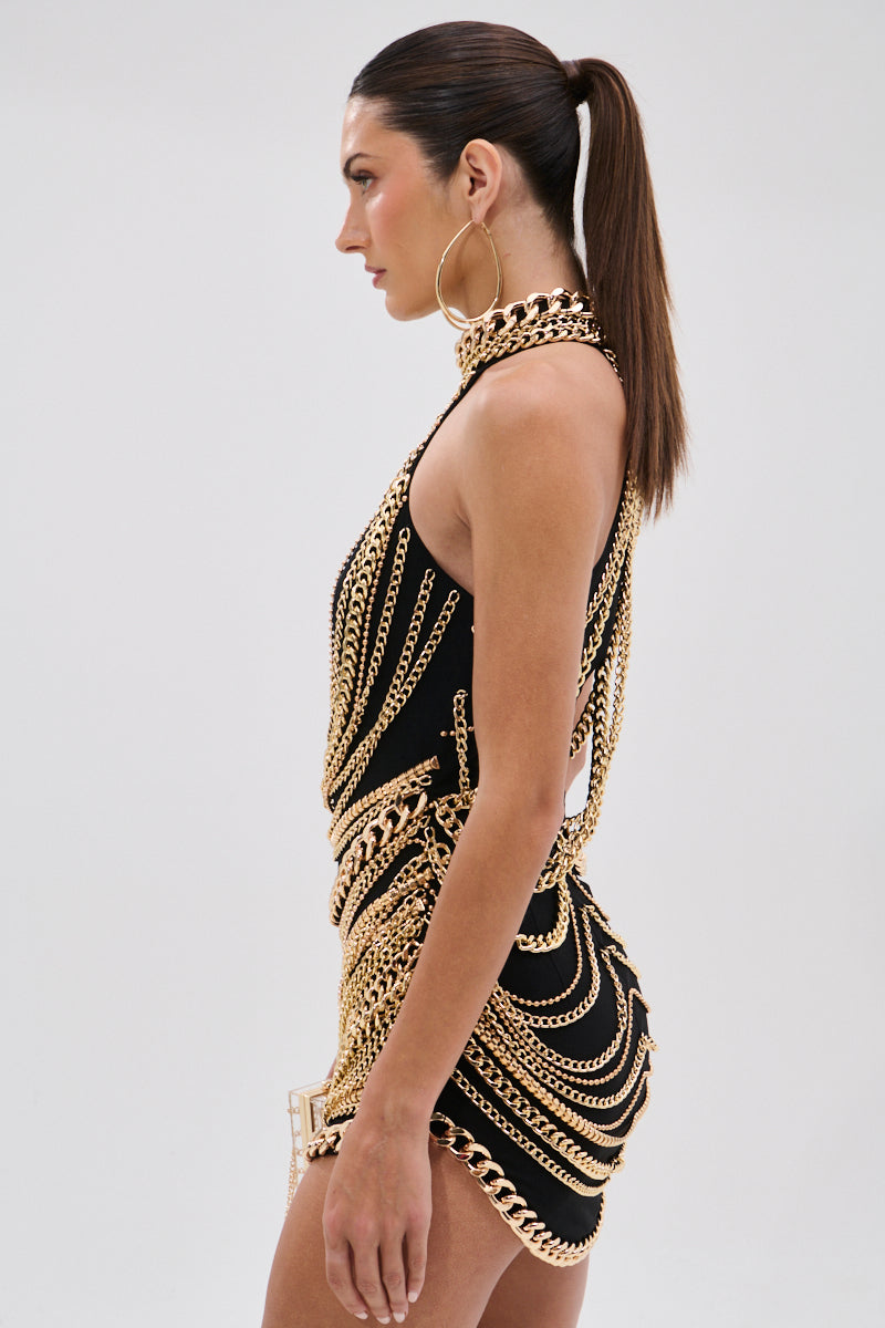 A woman with a sleek ponytail models the STAND OUT CHAIN MINI DRESS, a sleeveless black mesh mini dress with layered gold-tone chains and large hoop earrings, shown in profile against a plain light background.