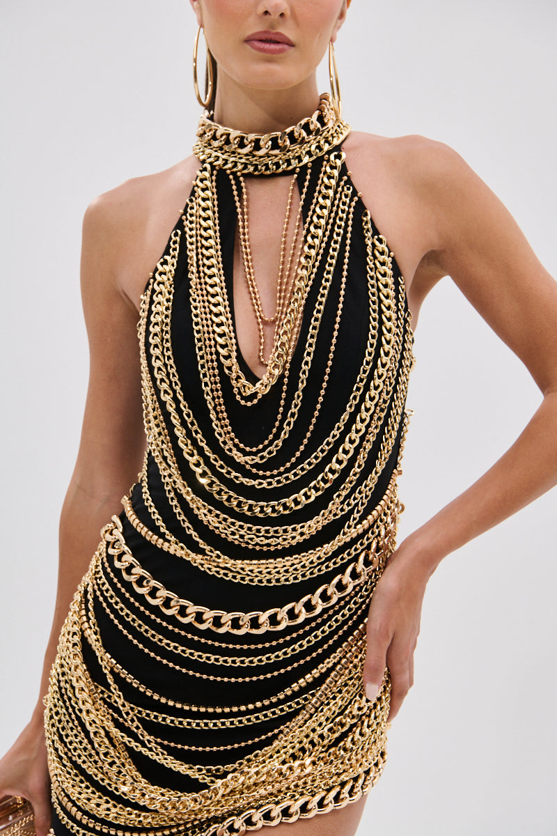A woman wears the STAND OUT CHAIN MINI DRESS, a sleeveless black mini dress with a high neckline, detailed with layered gold-tone chains, and accessorizes with large hoop earrings.