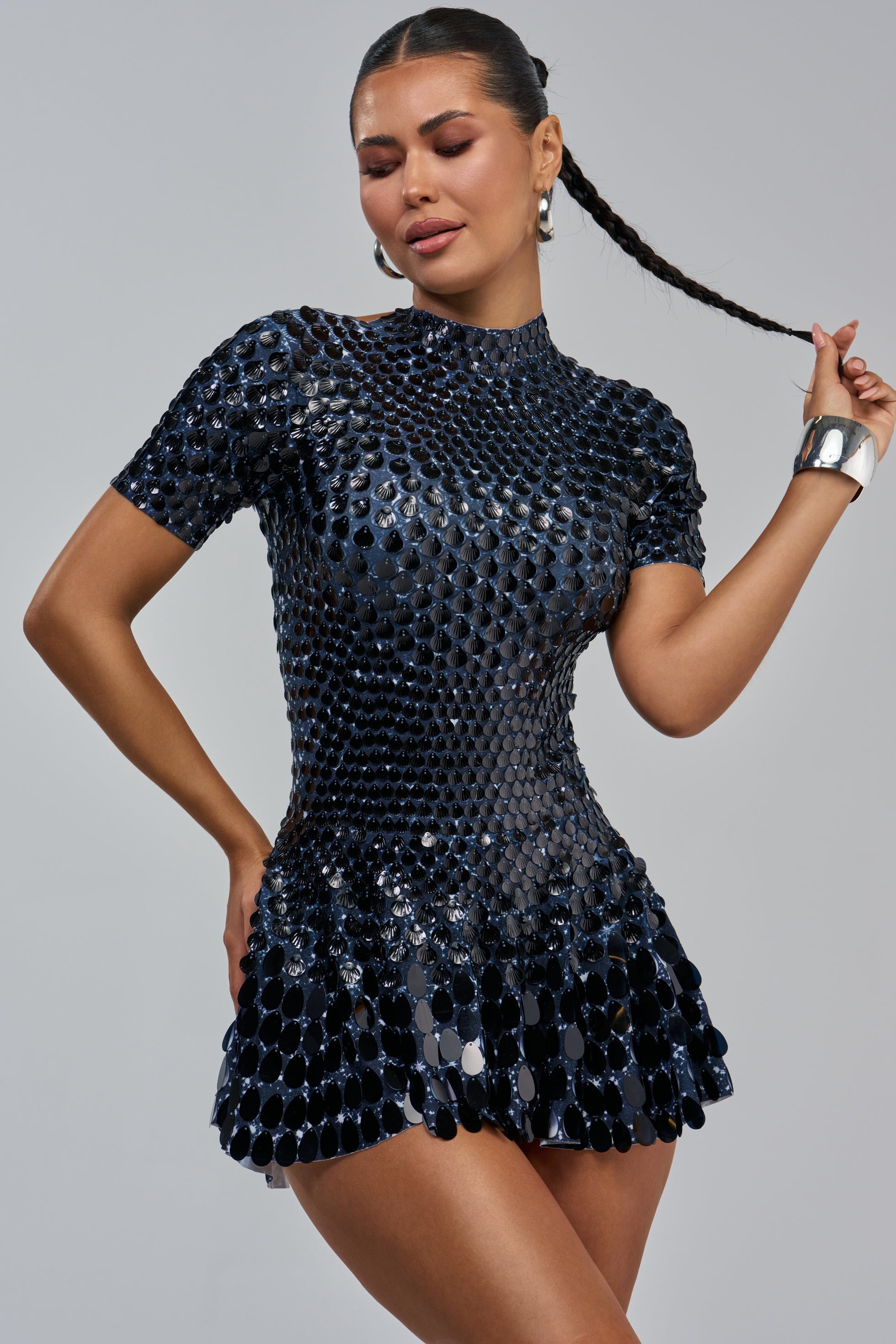 A woman poses confidently against a gray background in the FAR FROM BASIC SEQUIN MINI DRESS IN BLACK, featuring a short, shiny black design with metallic disc details. She accessorizes her look with hoop earrings, a wide silver bracelet, and holds her long braided hair.