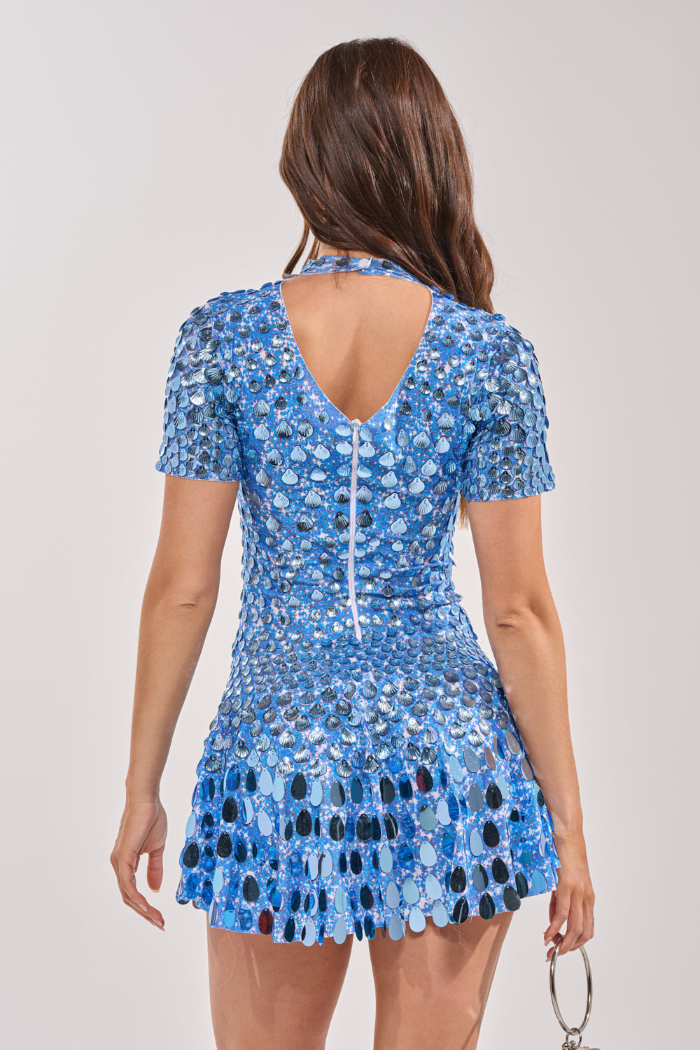 A woman with long brown hair is seen from the back, wearing the FAR FROM BASIC SEQUIN MINI DRESS IN BLUE with a keyhole cutout and a silver ring-shaped purse—an ideal choice for a seaside dinner. 4
