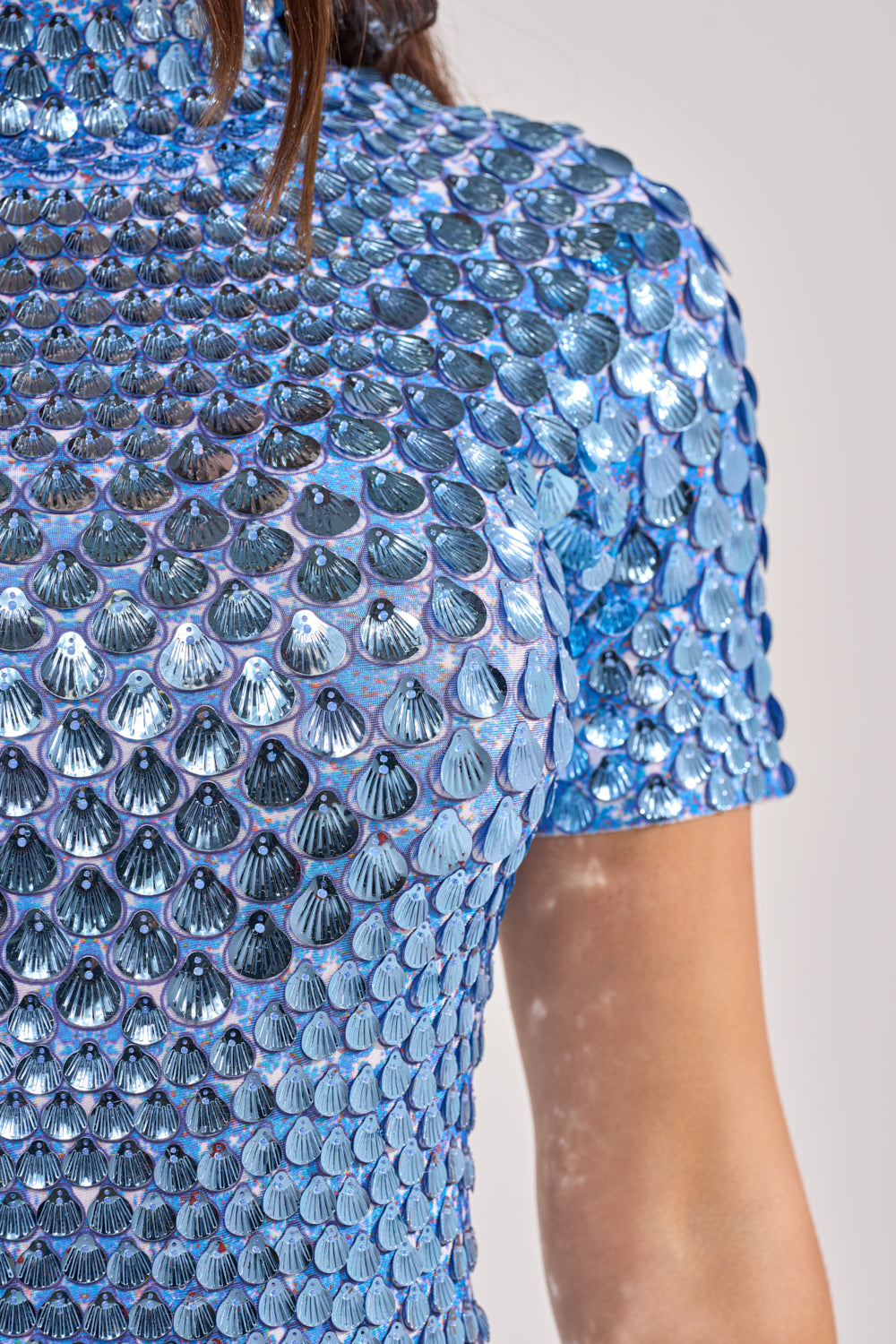 Close-up of a person wearing the FAR FROM BASIC SEQUIN MINI DRESS IN BLUE, featuring short sleeves and iridescent, overlapping sequin patterns. Only the upper torso and part of the arm are visible. 6