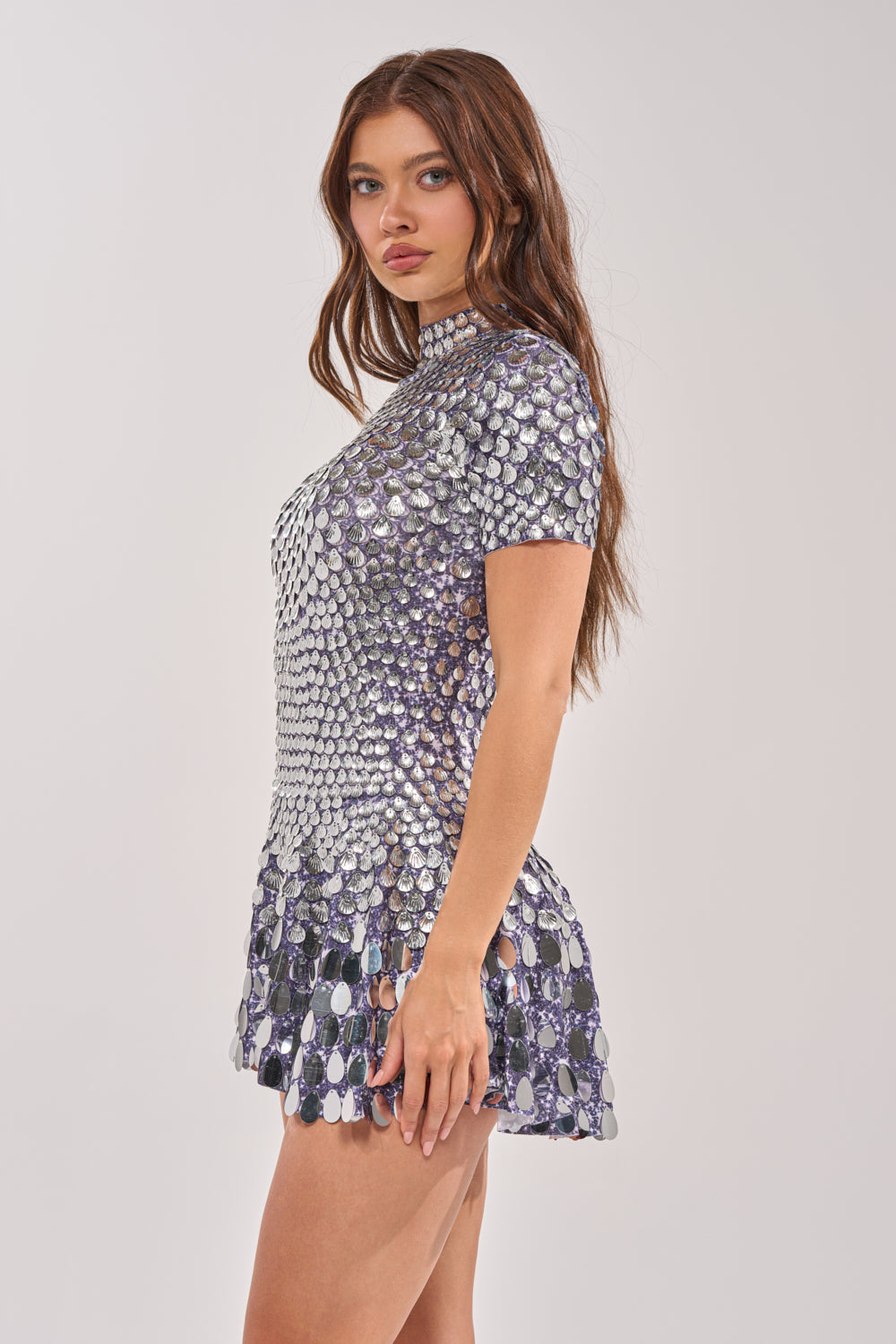 A young woman with long brown hair wears the FAR FROM BASIC SEQUIN MINI DRESS IN SILVER—short-sleeved, mock neck, and covered in silver sequins—standing against a plain light background and looking at the camera. 2