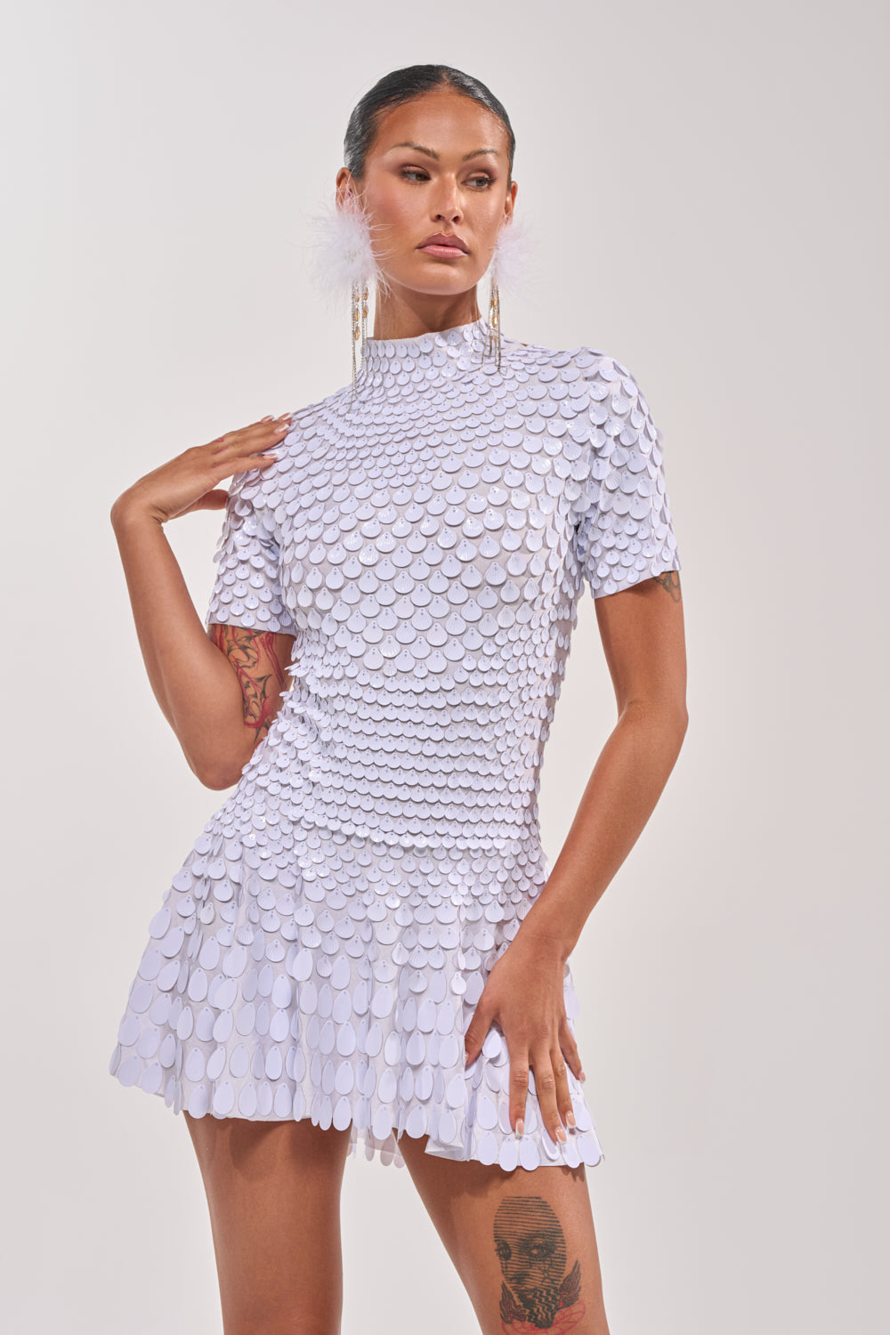 A person poses confidently in the FAR FROM BASIC SEQUIN MINI DRESS IN WHITE, styled with long earrings and feathered accessories, creating a chic beach party look against a plain light background. 0