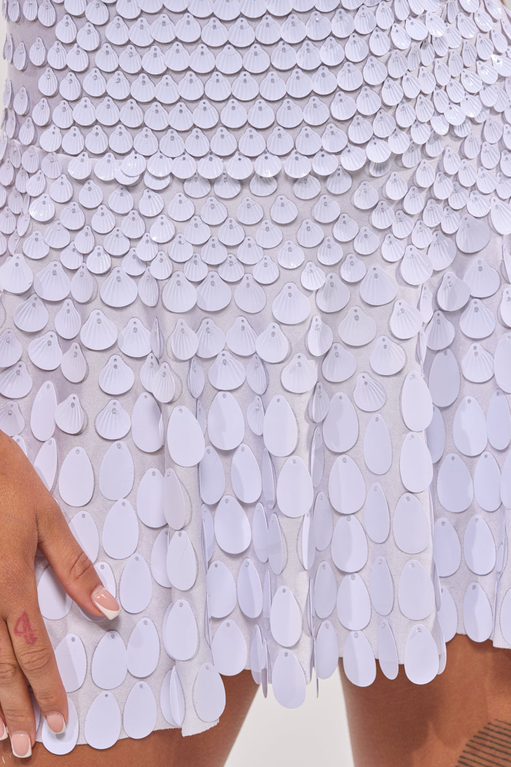Close-up of a person wearing the FAR FROM BASIC SEQUIN MINI DRESS IN WHITE, featuring layered petal-shaped sequins. One hand rests by their side, revealing light skin and a small red heart tattoo on the finger. 9